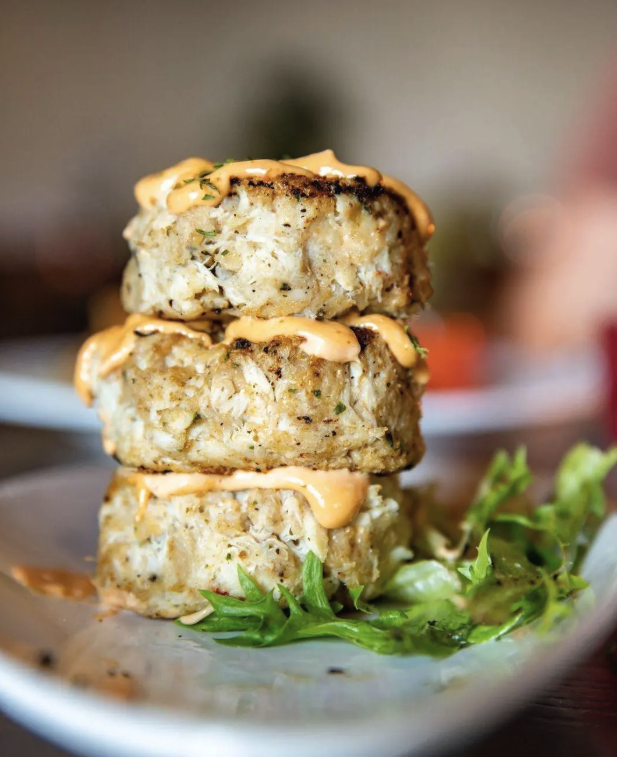 Stack of three crab cakes drizzled with sauce, served on a bed of lettuce on a white plate.