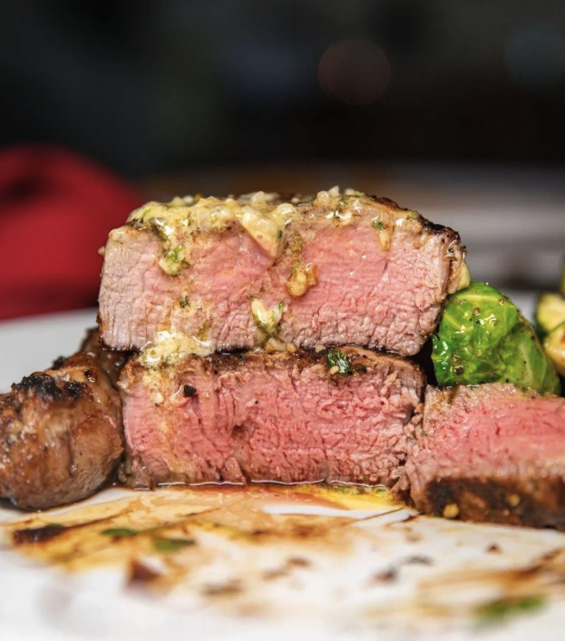Two thick-cut slices of medium-rare steak topped with herb butter, served with grilled vegetables and a sauce on the plate.