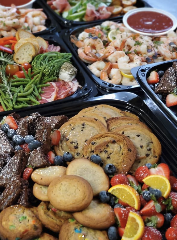 Assorted dishes including shrimp, pasta, vegetables, chocolate chip cookies, assorted berries, and sliced fruits, all in black serving trays for a buffet meal.