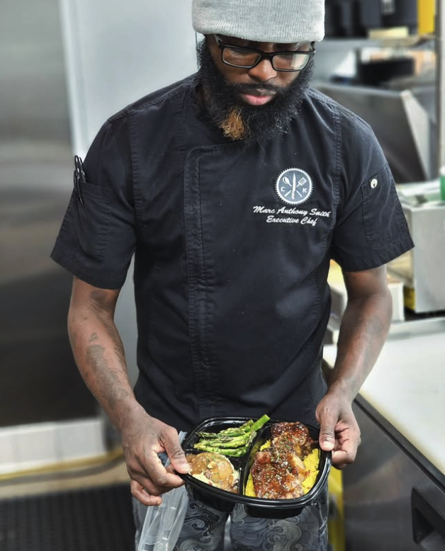 A man wearing a gray beanie, glasses, a black chef's uniform with an embroidered logo and text, is holding a black takeout container with meat and vegetables. The background shows a commercial kitchen with stainless steel appliances.