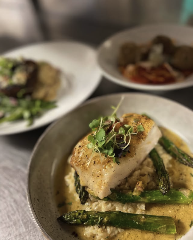 A plated dish featuring a piece of cooked white fish garnished with microgreens, served over asparagus and rice with a creamy sauce. Two other plates of food are partially visible in the background.