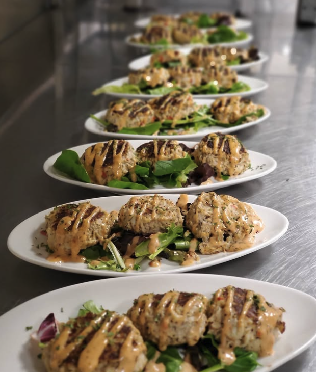 A row of white plates with food, each featuring three chicken meatballs drizzled with sauce on a bed of greens, on a stainless steel countertop.