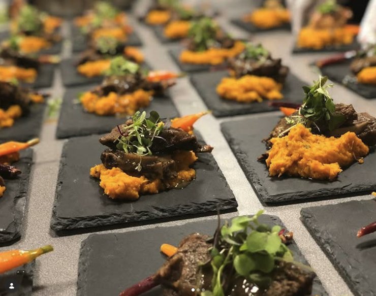 Multiple small servings of gourmet appetizers on black slate plates, featuring mashed sweet potatoes, slow-cooked beef, microgreens, and roasted peppers.