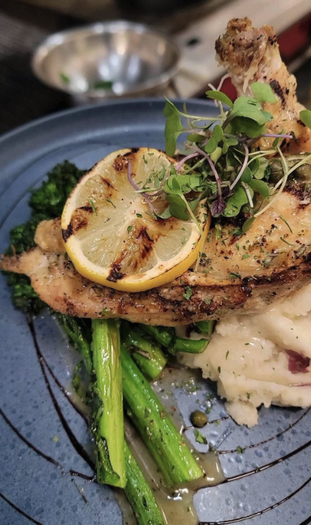Grilled chicken breast topped with lemon slices and microgreens, served with sautéed asparagus, broccoli, and mashed potatoes.