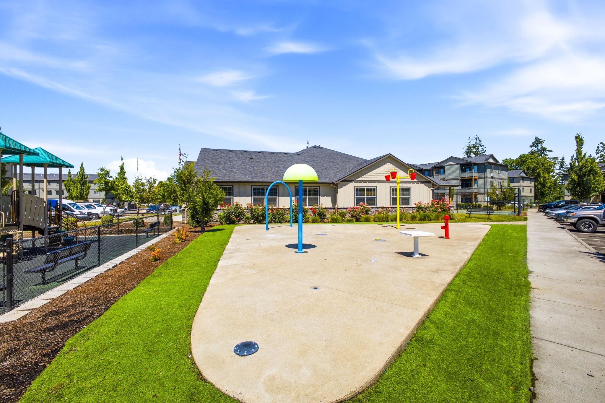 Community park with a concrete trail, a playground with a mushroom-shaped shade, sports equipment, and a residential building in the background under a blue sky.