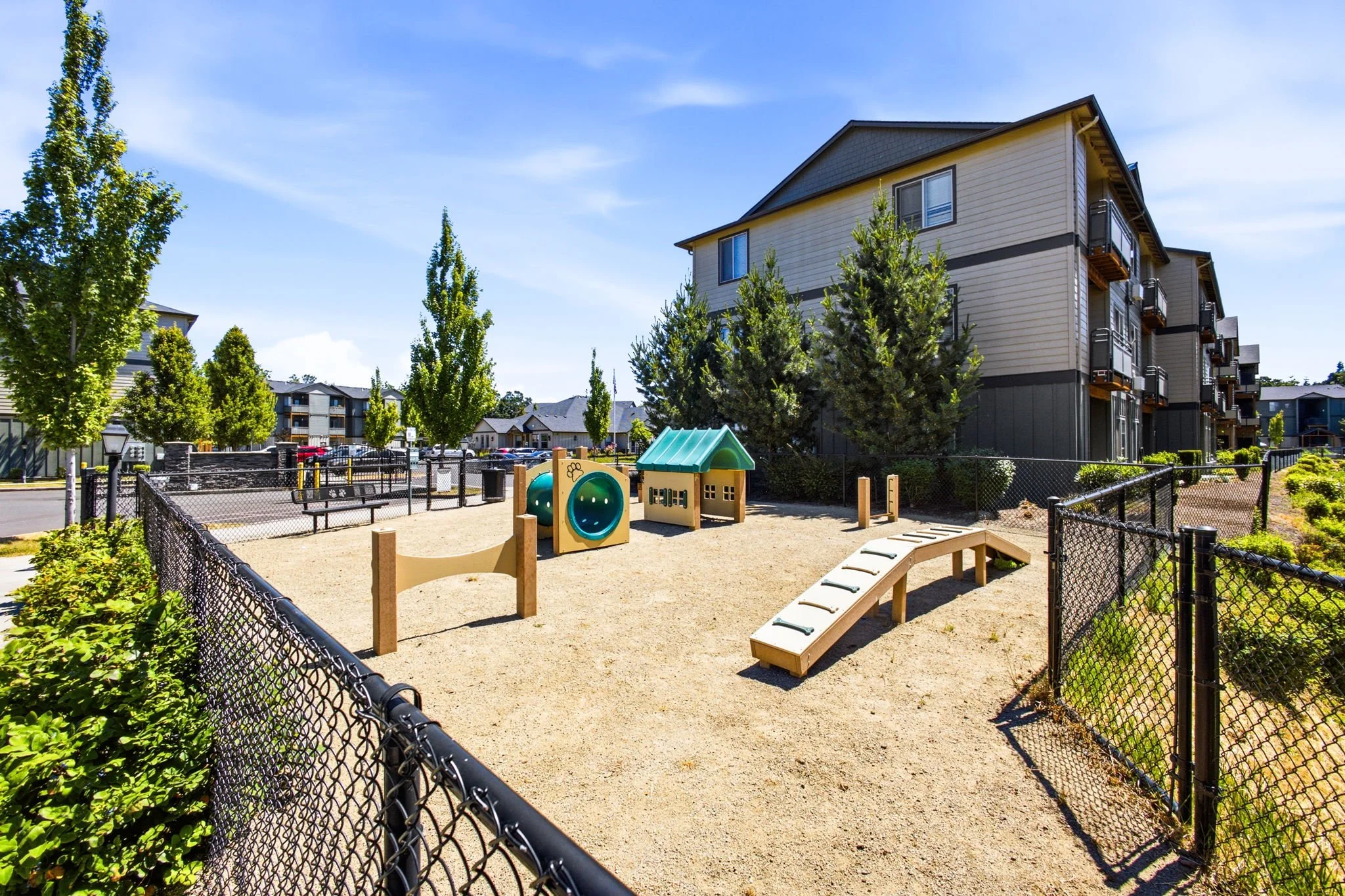 An outdoor playground area surrounded by a black chain-link fence, with a small playhouse, a slide, and a sandbox, located near multi-story residential buildings with beige siding and balconies. There are trees and parking lots visible in the background under a clear blue sky.