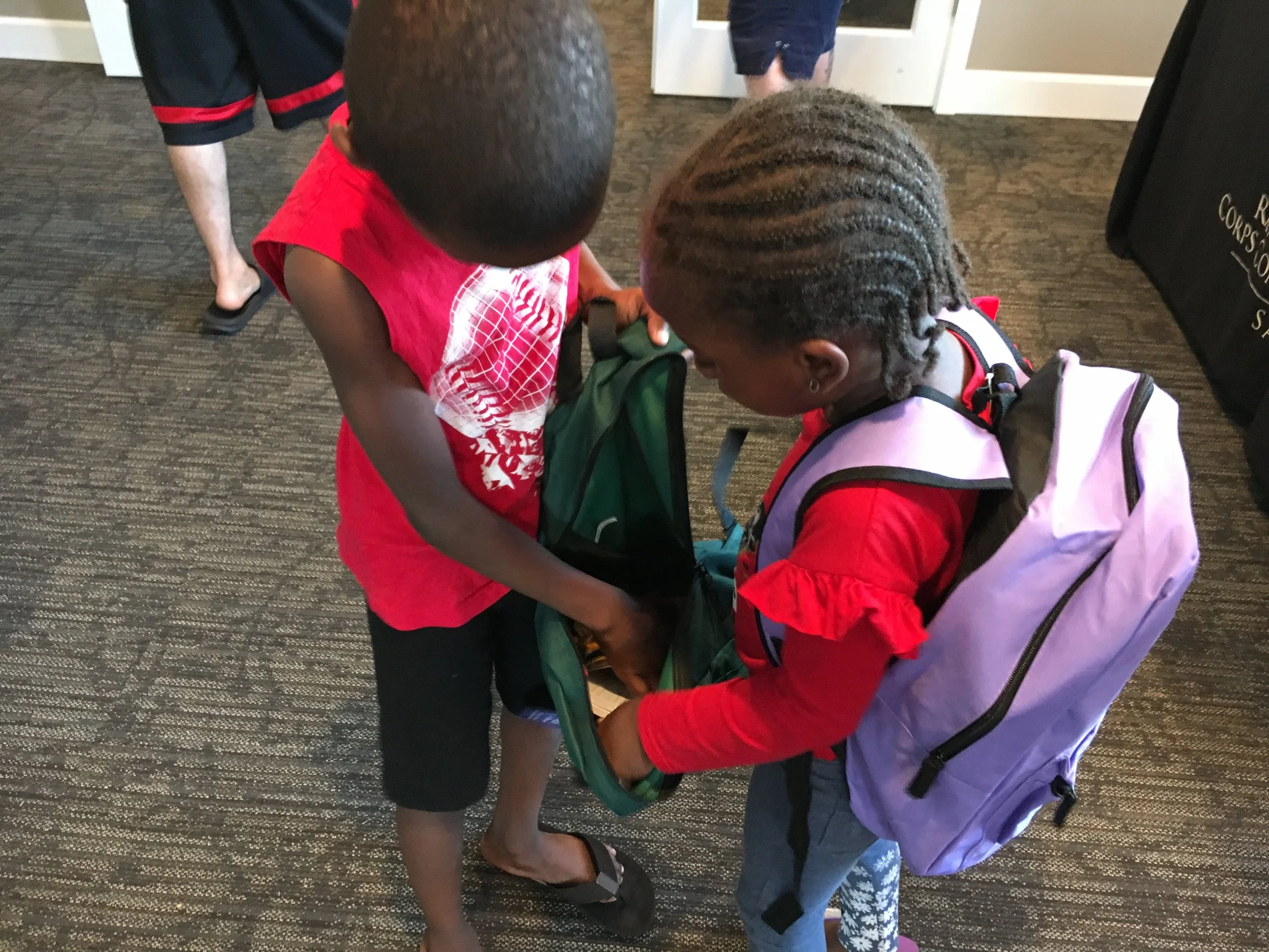 Two children are looking into an open backpack. One child has short hair and is wearing a red dress, while the other has braided hair and is wearing a pink jacket and backpack. They are standing on a carpeted floor near a door.