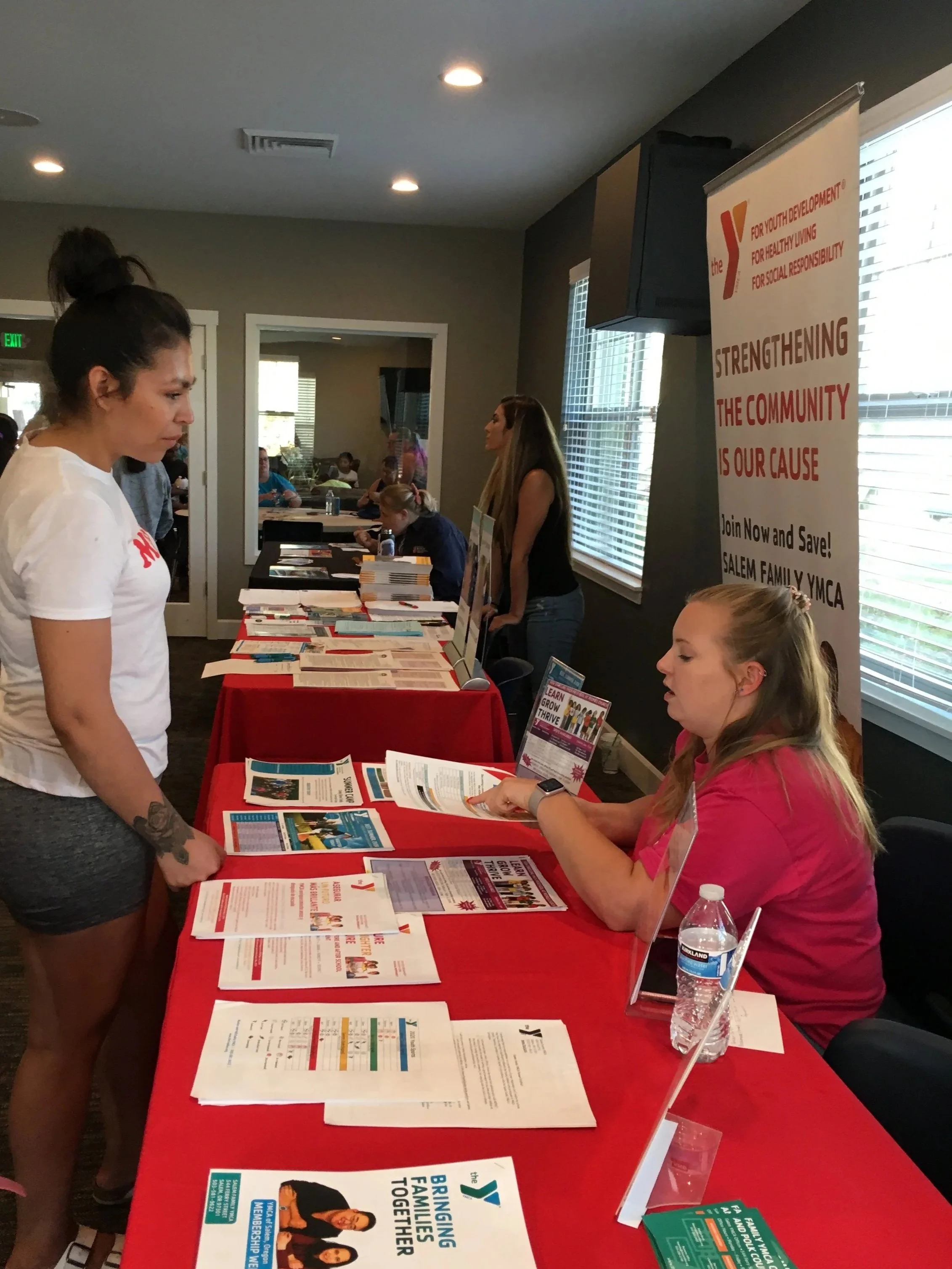 A woman is standing and talking to a seated woman at an event booth with informational brochures and pamphlets about the YMCA's programs, with a large banner in the background promoting community strengthening, youth development, and social responsibility.