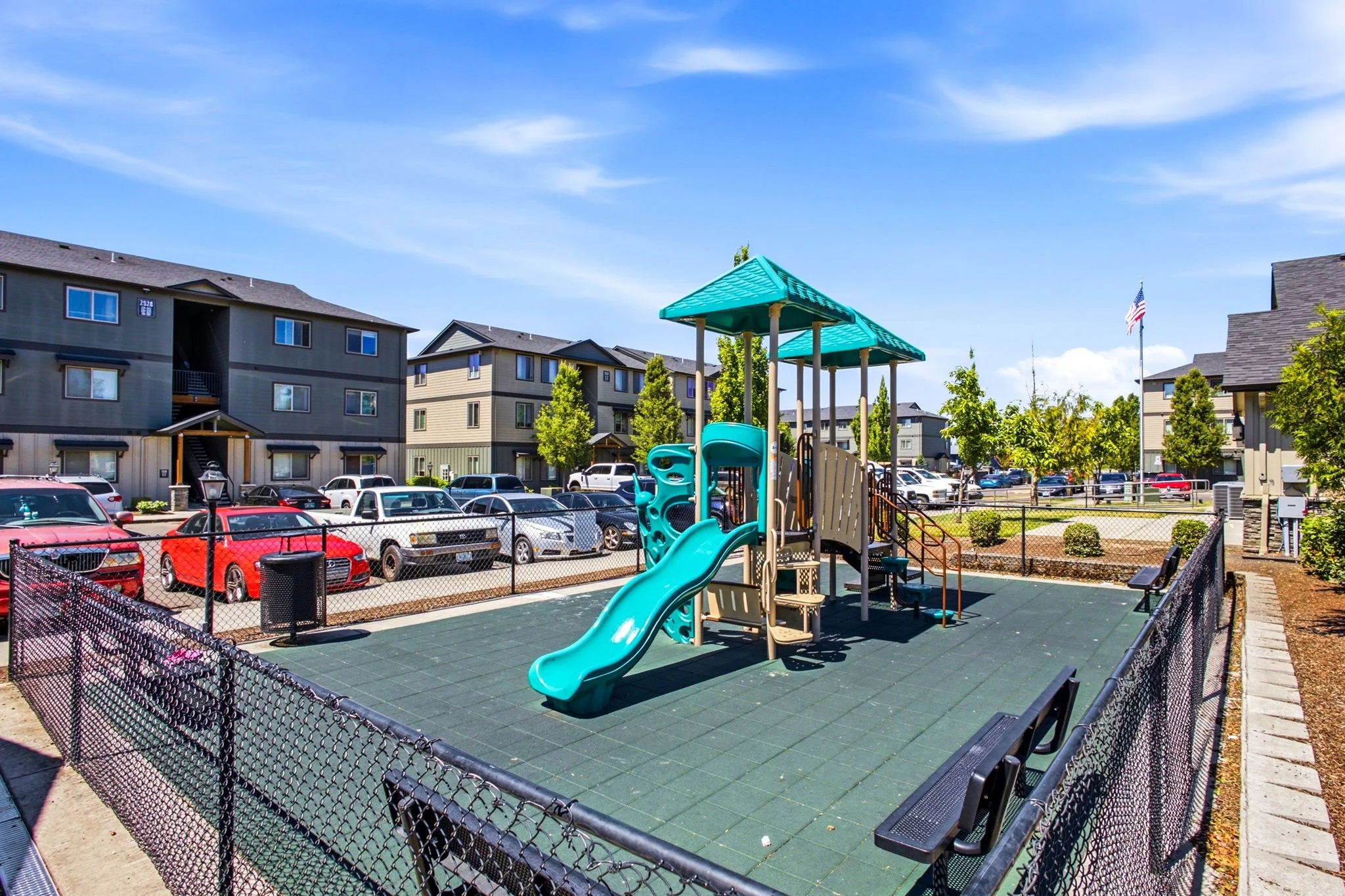 Playground with blue slide and shade structures surrounded by a black fence, with parking lot and multi-story apartment buildings in the background.