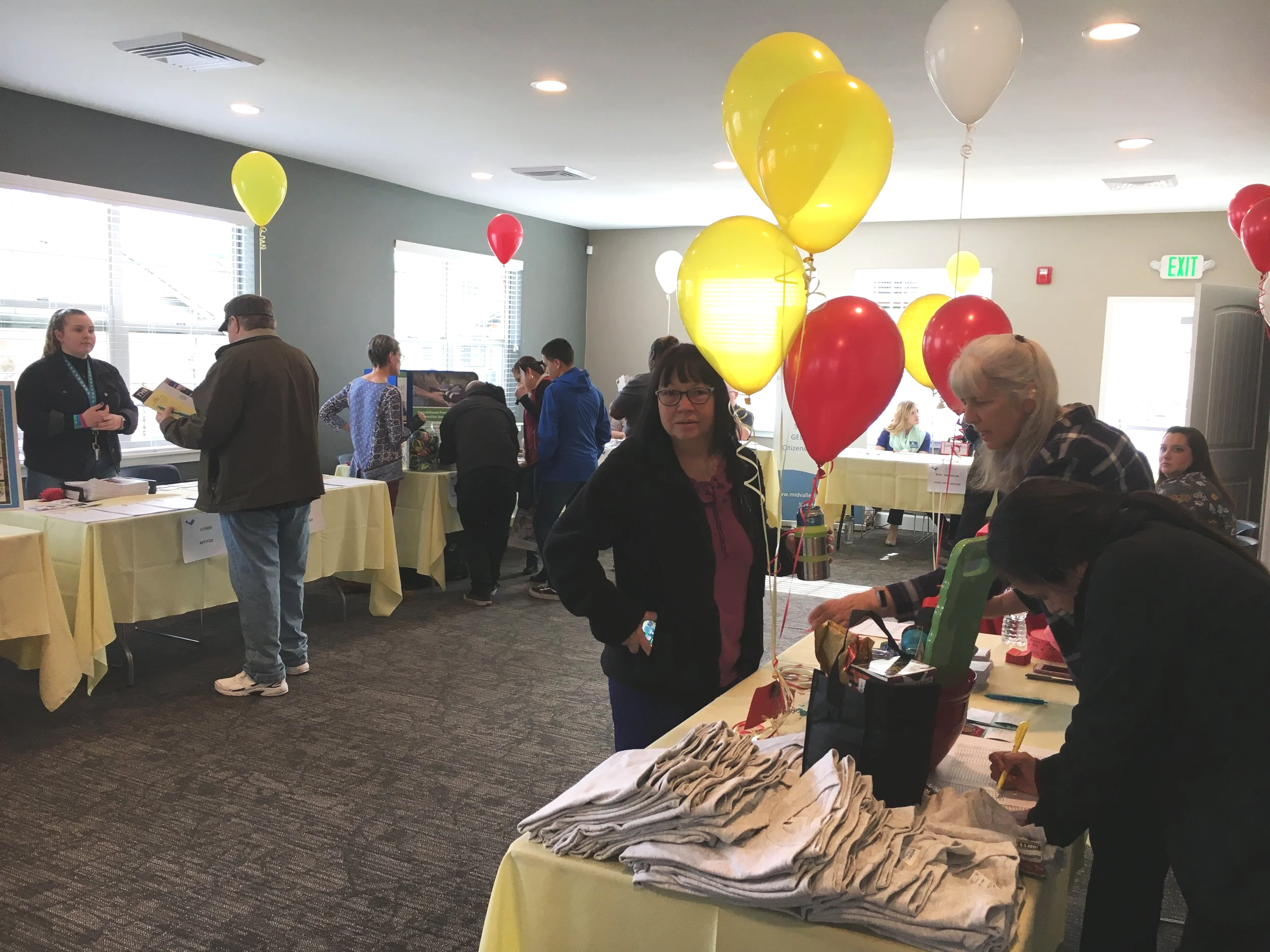 An indoor event with balloons, tables, and people engaging with various booths, including a registration or information desk.
