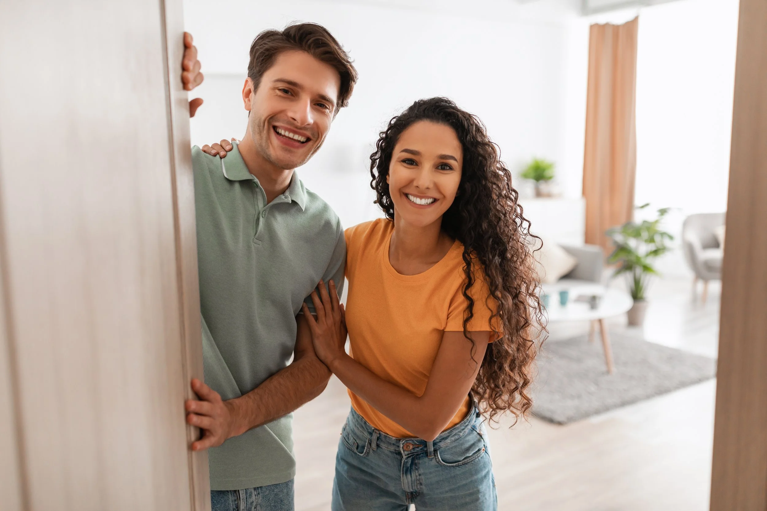A smiling young man and woman peering from behind a door, with the man slightly what and the woman with long, curly hair, in a bright living room with plants and furniture.