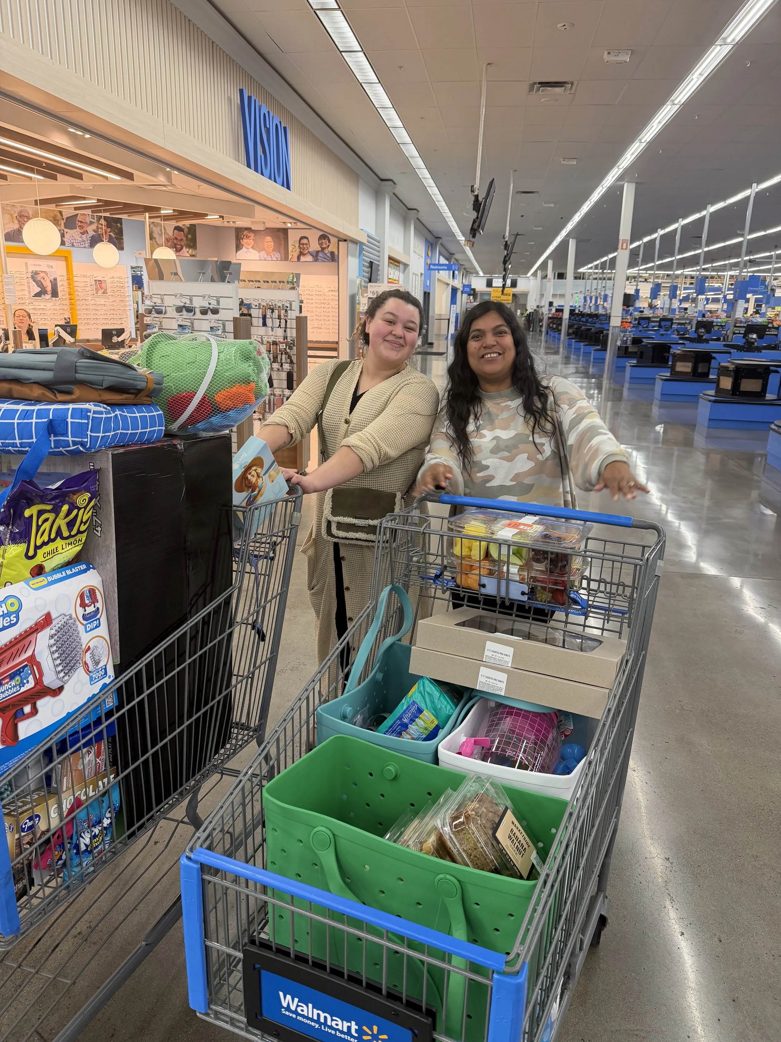 Two women smiling with shopping carts filled with groceries at a Walmart store, next to a store entrance with a 'Vision' sign and checkout counters in the background.