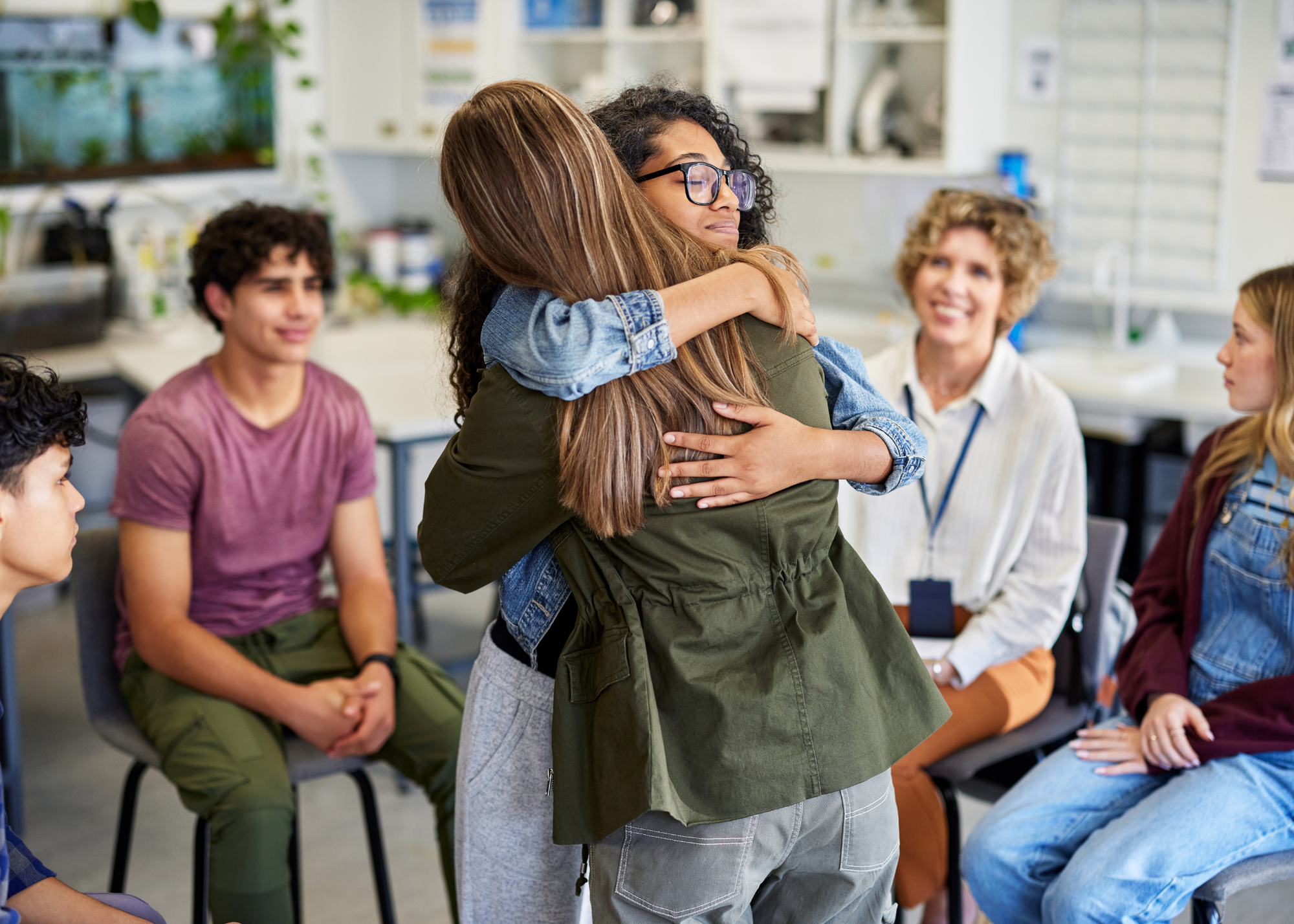 Two women hugging each other in front of a seated group of people in a classroom or seminar room. The women appear to be sharing an emotional moment while others watch with smiles.