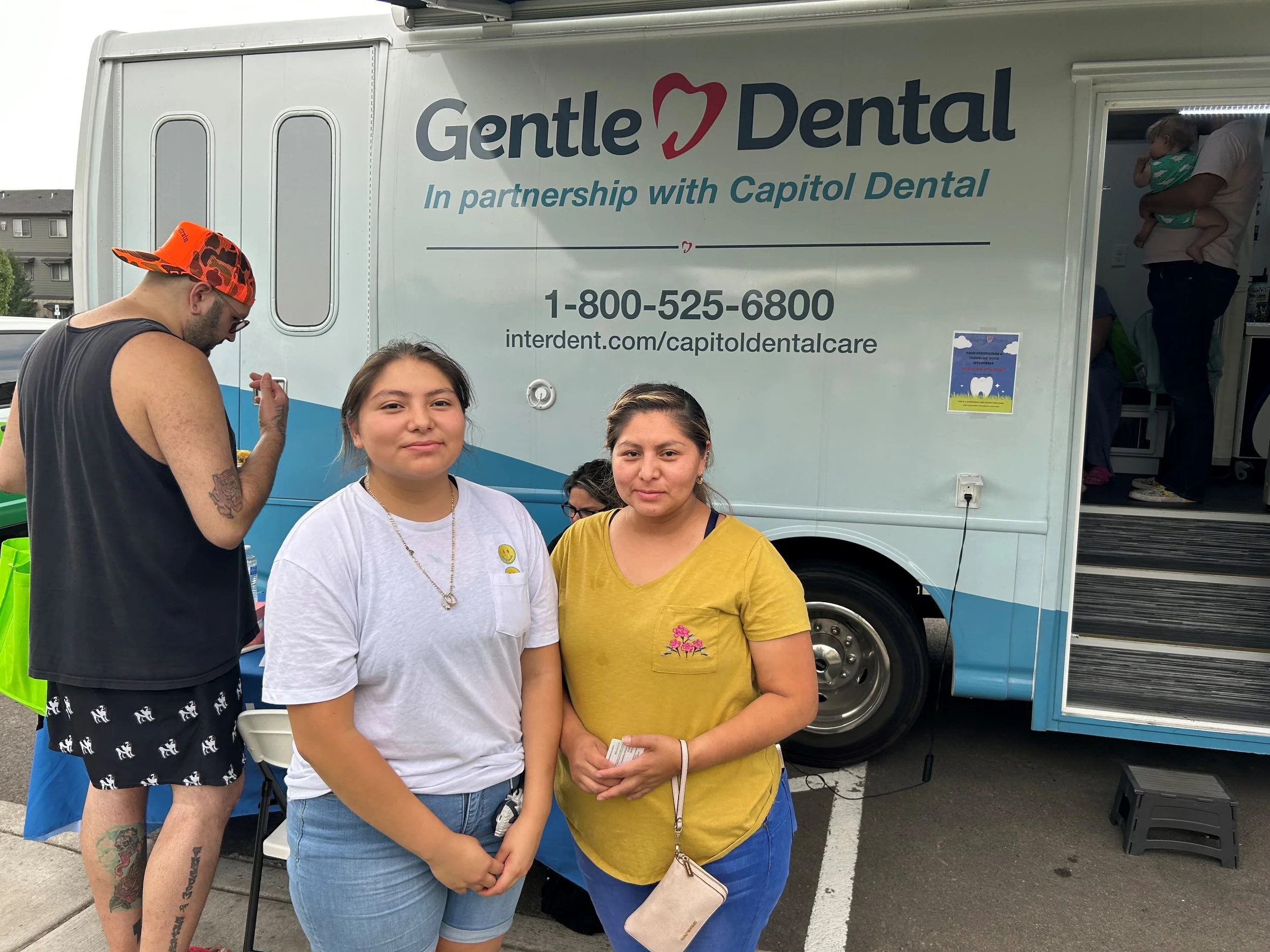 Three women standing in front of a mobile dental clinic, with two women and a man inside the clinic. The mobile clinic has a sign that reads 'Gentle Dental in partnership with Capitol Dental,' with a phone number and website. One woman is wearing a white shirt with yellow smiley face button, another wearing a yellow shirt and carrying a small beige bag, and the third woman is wearing a black shirt and shorts with a cat pattern, topped with an orange cap.