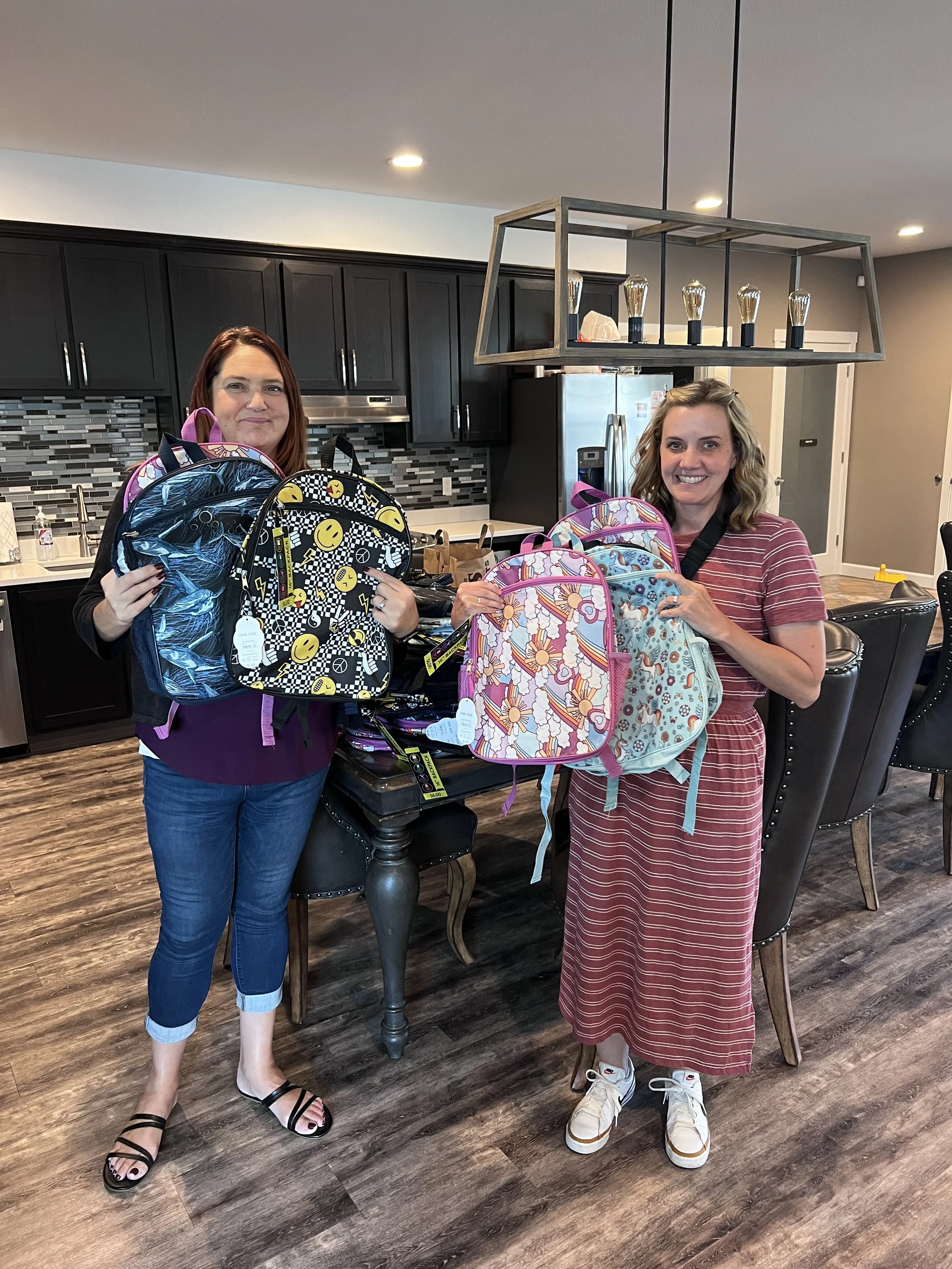 Two women in a kitchen each hold backpacks with colorful patterns, smiling at the camera.