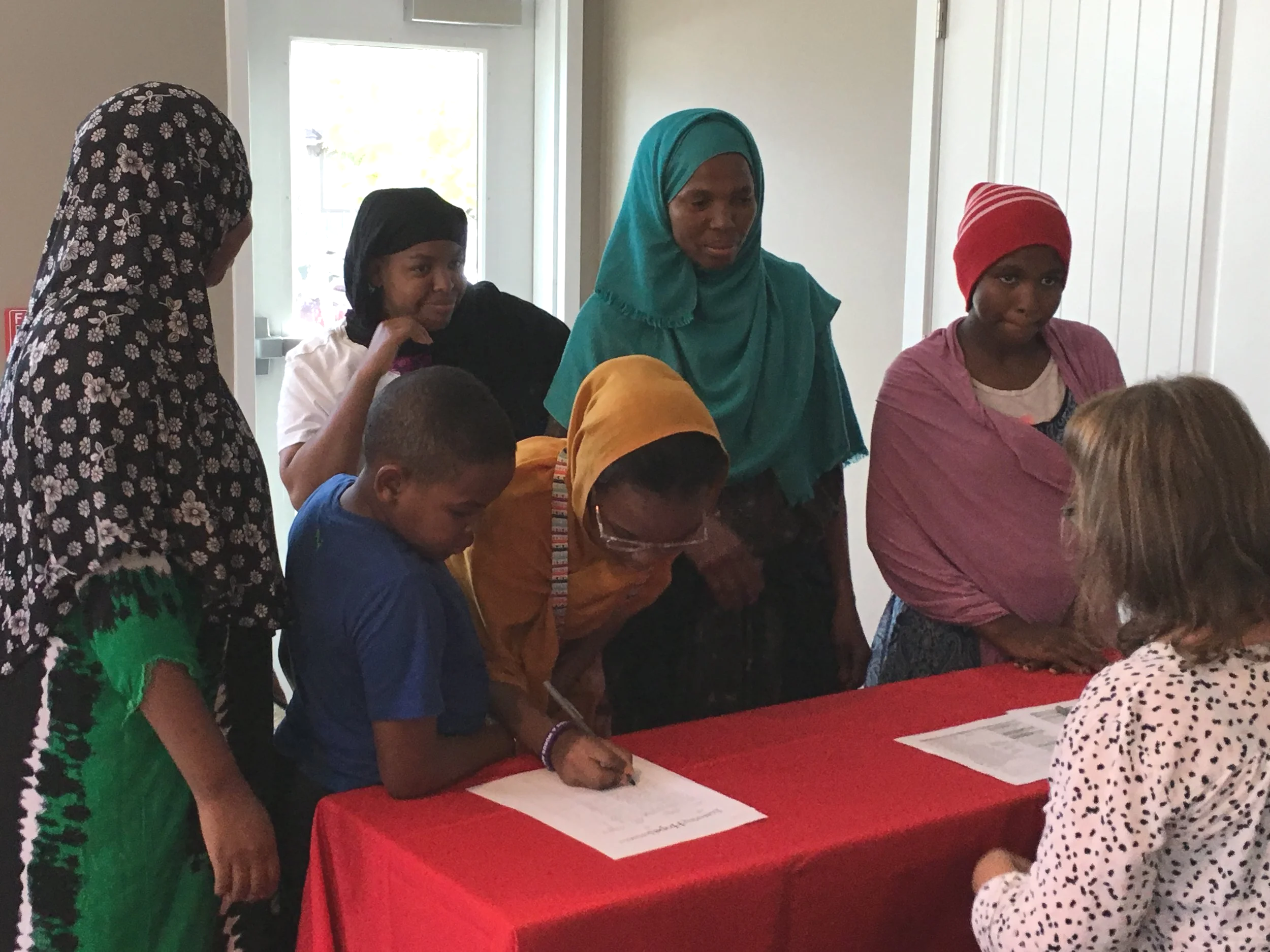 A group of women and children gathered around a table with documents, some women are writing or reading.