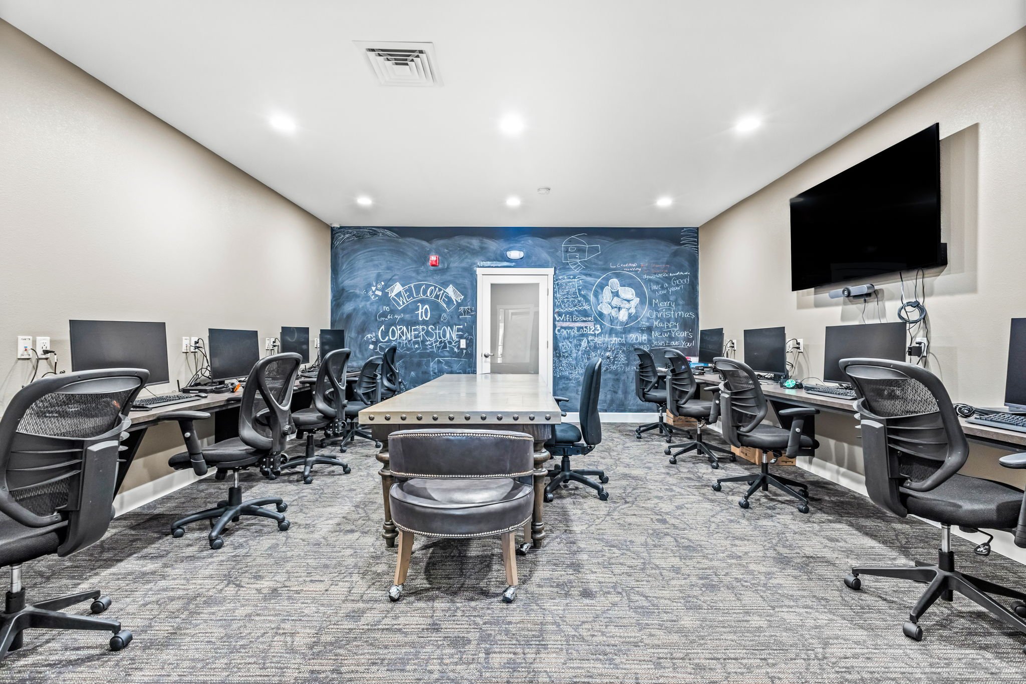 A computer classroom with multiple desks and chairs arranged in a U-shape around a central table. Each desk has a monitor, keyboard, and mouse. The room features a chalkboard wall with handwritten notes and drawings, including a welcome message, holiday greetings, and various sketches. A large flat-screen TV is mounted on the right wall.