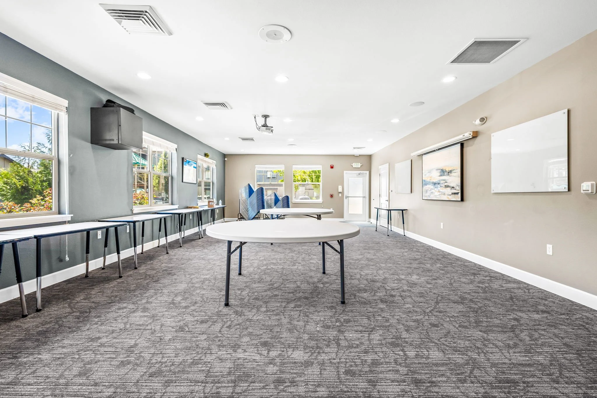 A spacious conference room with gray walls and multiple windows with blinds, decorated with abstract artwork, and furnished with tables and blue chairs stacked in the corner.