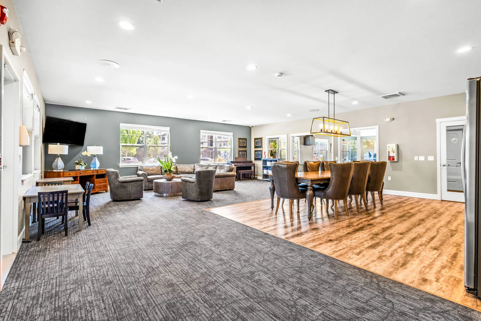 Open-concept living and dining room with beige and gray walls, large windows, gray sofa, wooden dining table with high-back chairs, and a mix of carpeting and hardwood flooring.