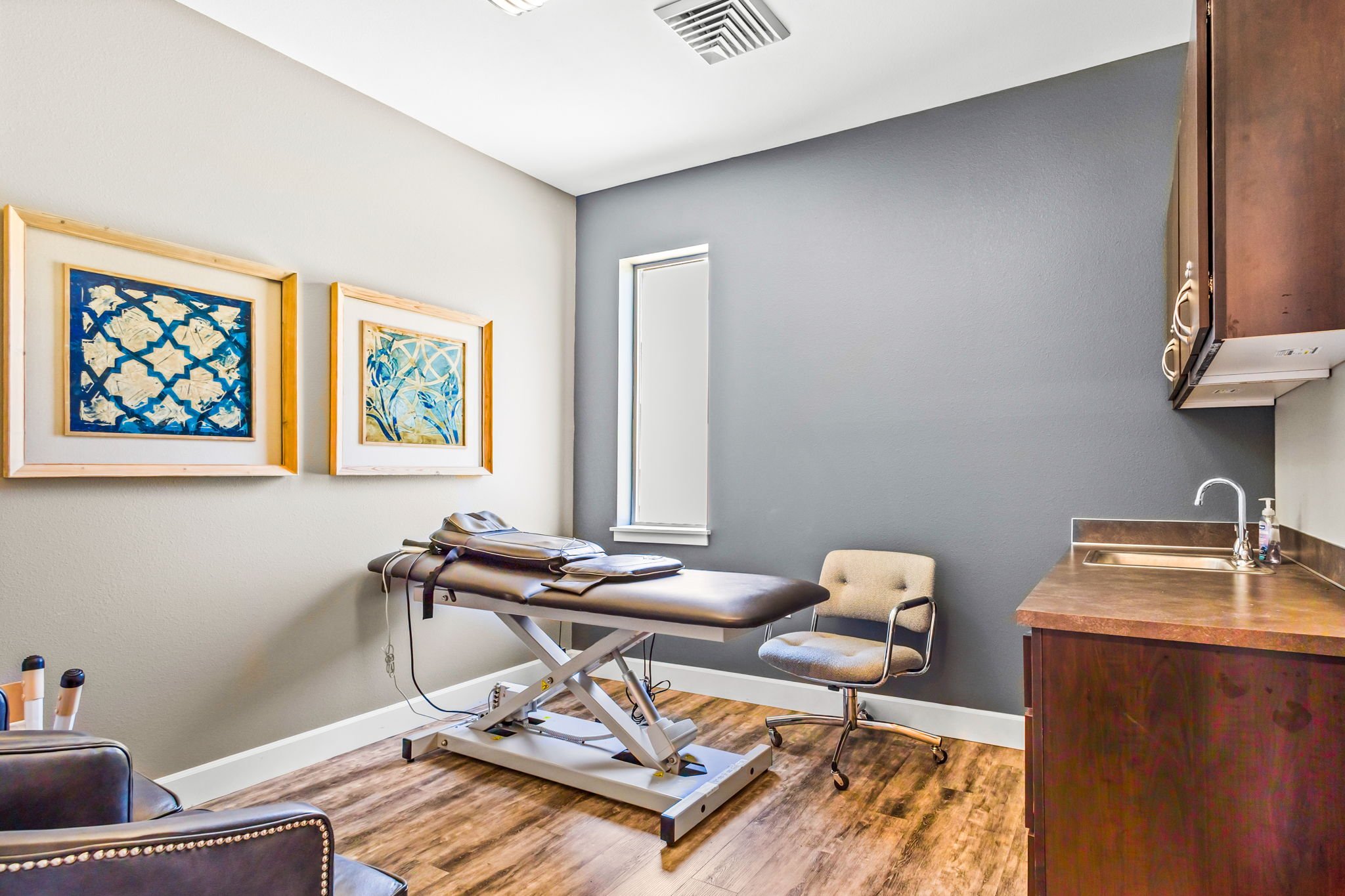 Medical examination room with an examination table, chairs, framed artwork on the wall, a window, and a wooden cabinet, in a healthcare facility.