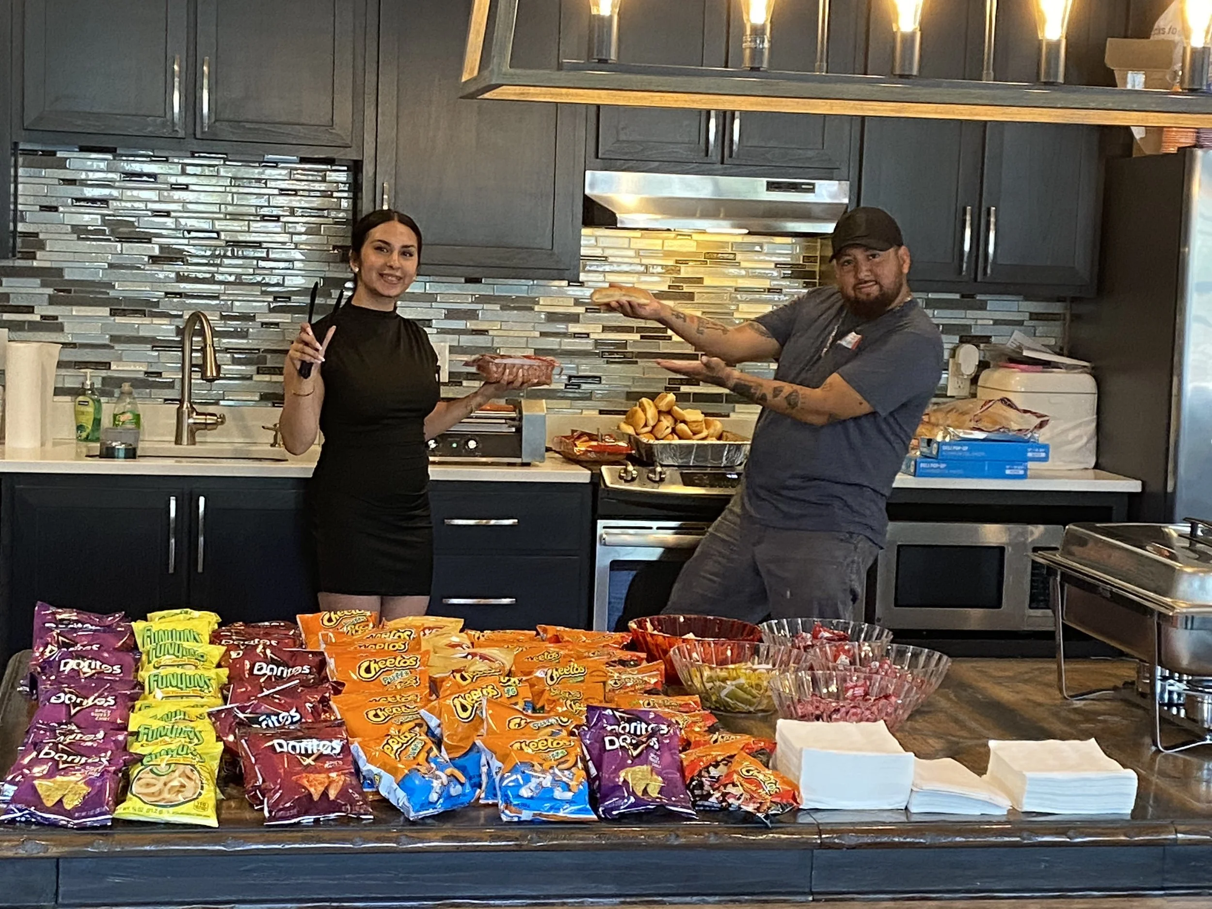 Two people standing behind a table filled with various snack foods, with bags of chips in the foreground. The woman is holding tongs and smiling, the man is gesturing with his hands, in a kitchen with dark cabinets and a mosaic tile backsplash.