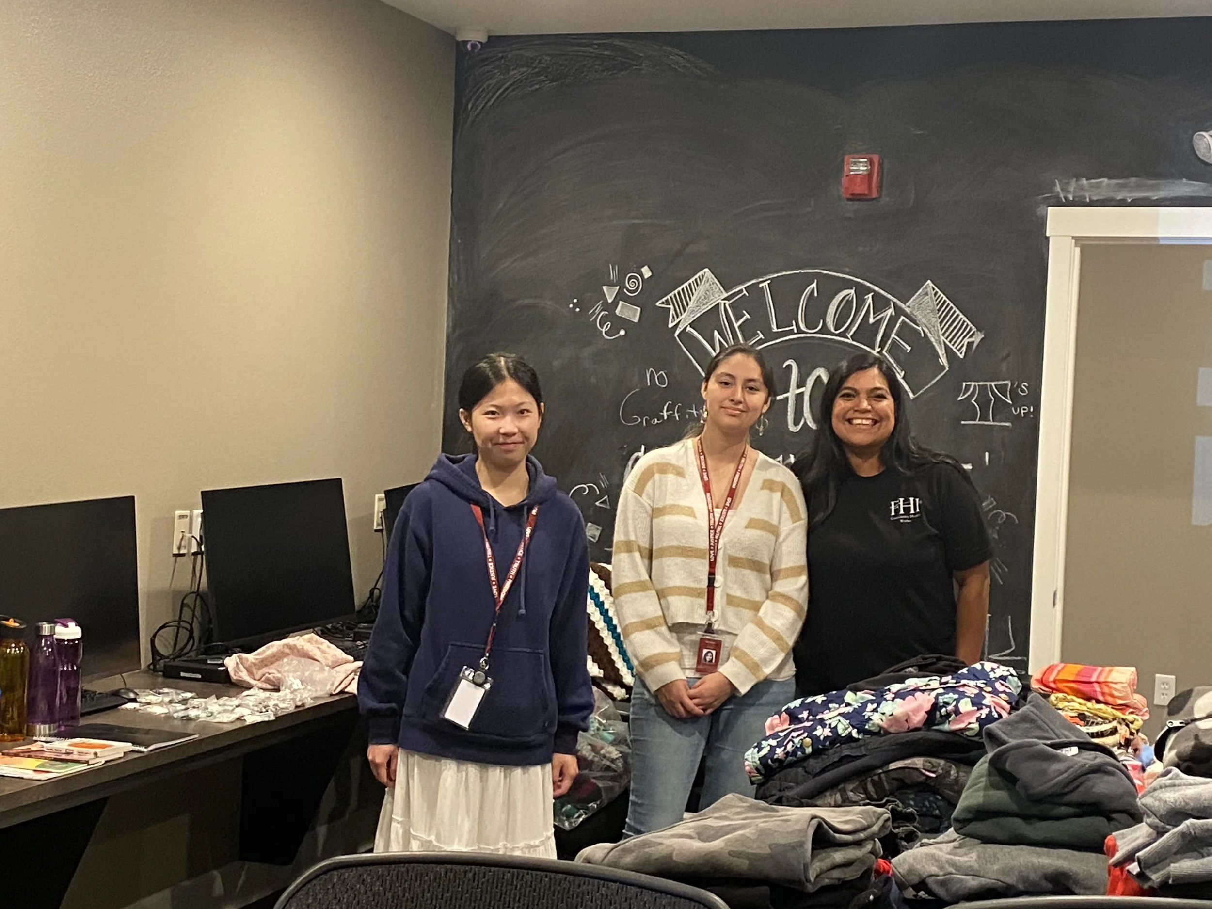 Three women standing behind a table with clothing, fabric, and accessories on it, in a room with a black chalkboard wall decorated with an illustration and the word "WELCOME".