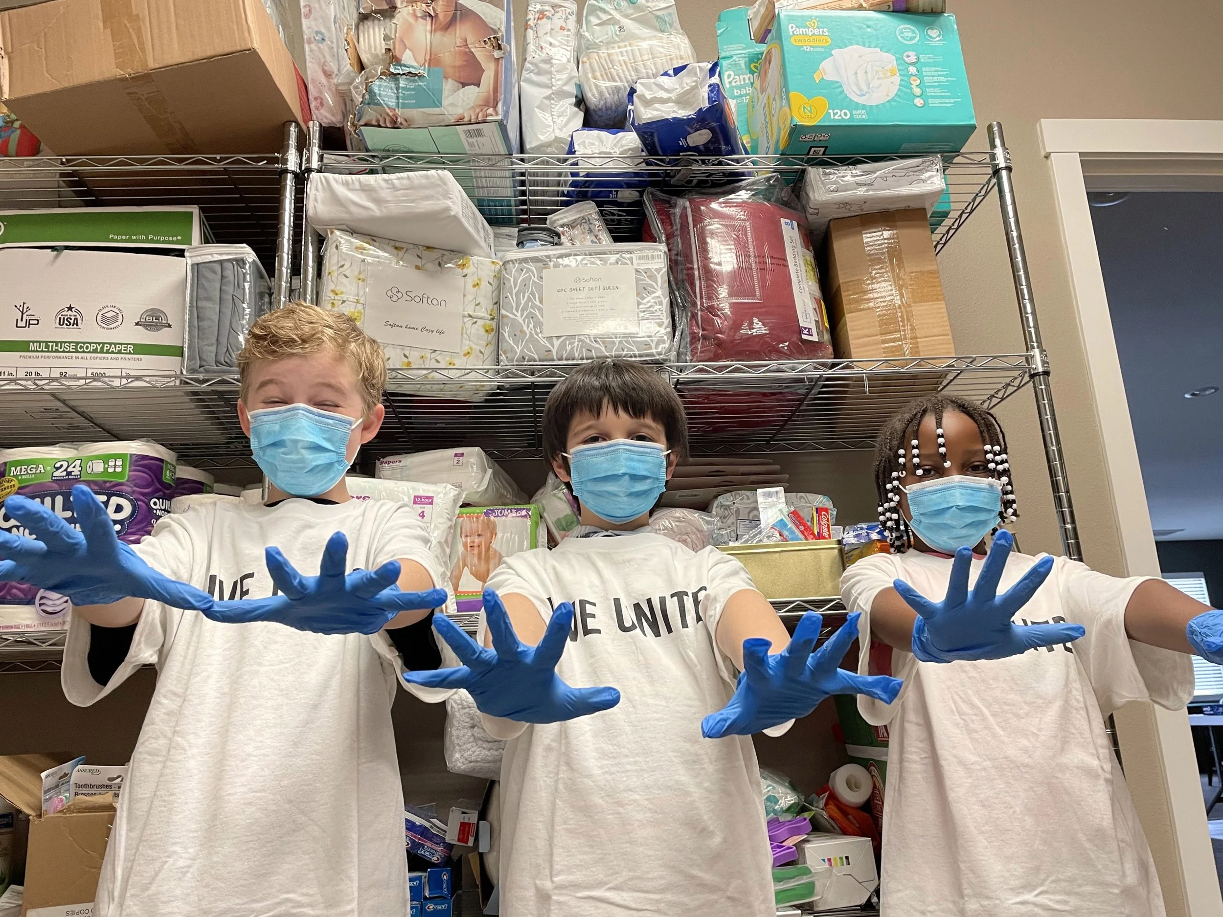 Three children wearing protective face masks and blue gloves standing in front of store shelves filled with household and baby supplies, smiling and posing for the camera.