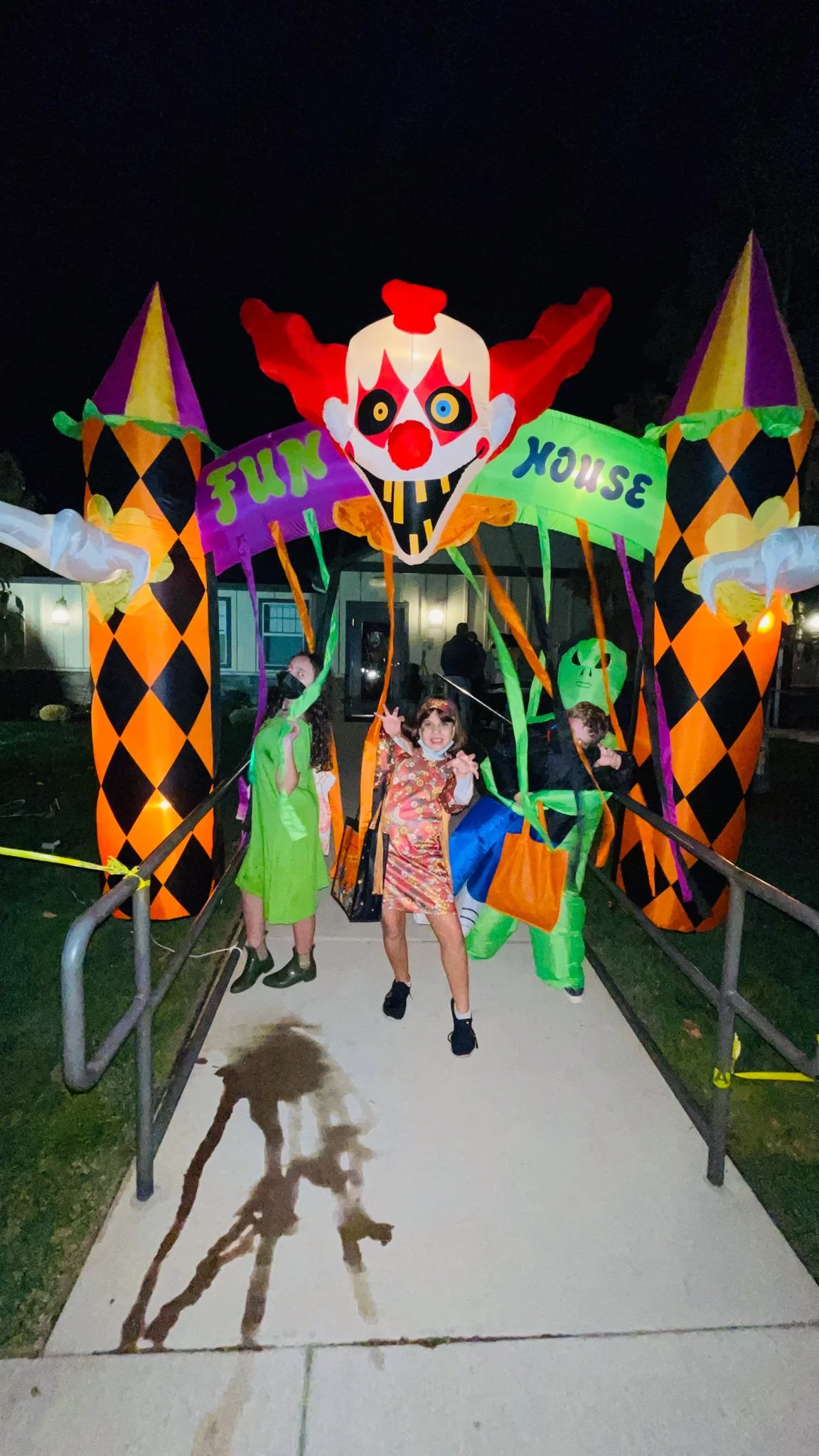 People dressed in Halloween costumes standing in front of a large, illuminated clown-themed Halloween decoration at night. The decoration features a smiling clown face with orange and purple pointed hats and a banner that reads 'FUN HOUSE'.