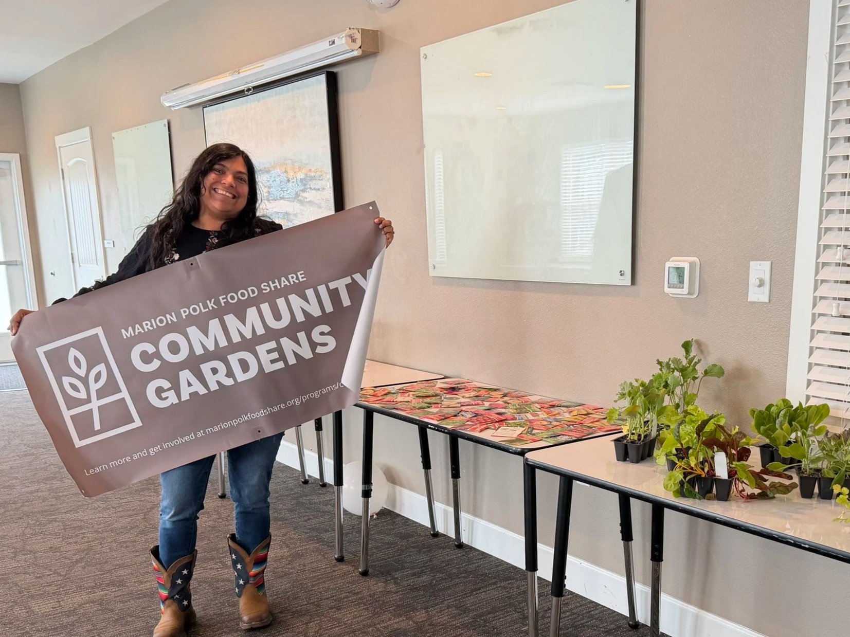 Woman smiling and holding a Banner that says 'Marion Polk Food Share Community Gardens' in an indoor space with a table of seedlings and potted plants, and whiteboards on the wall.