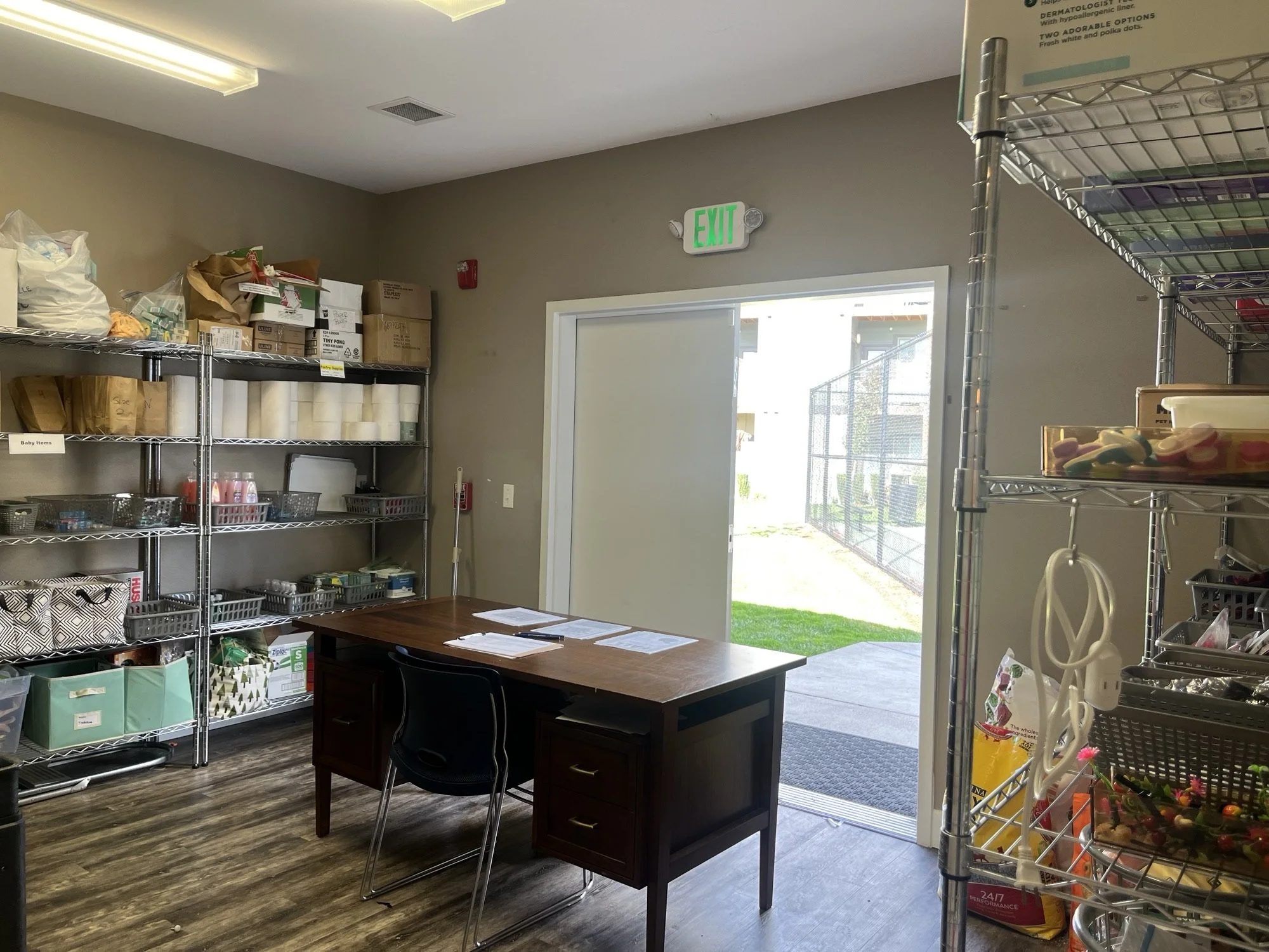 Interior of a store or office with metal shelving, a wooden desk, and an open door leading outside. Shelves contain various boxes, supplies, and toys. Fluorescent lights and a green exit sign are visible.