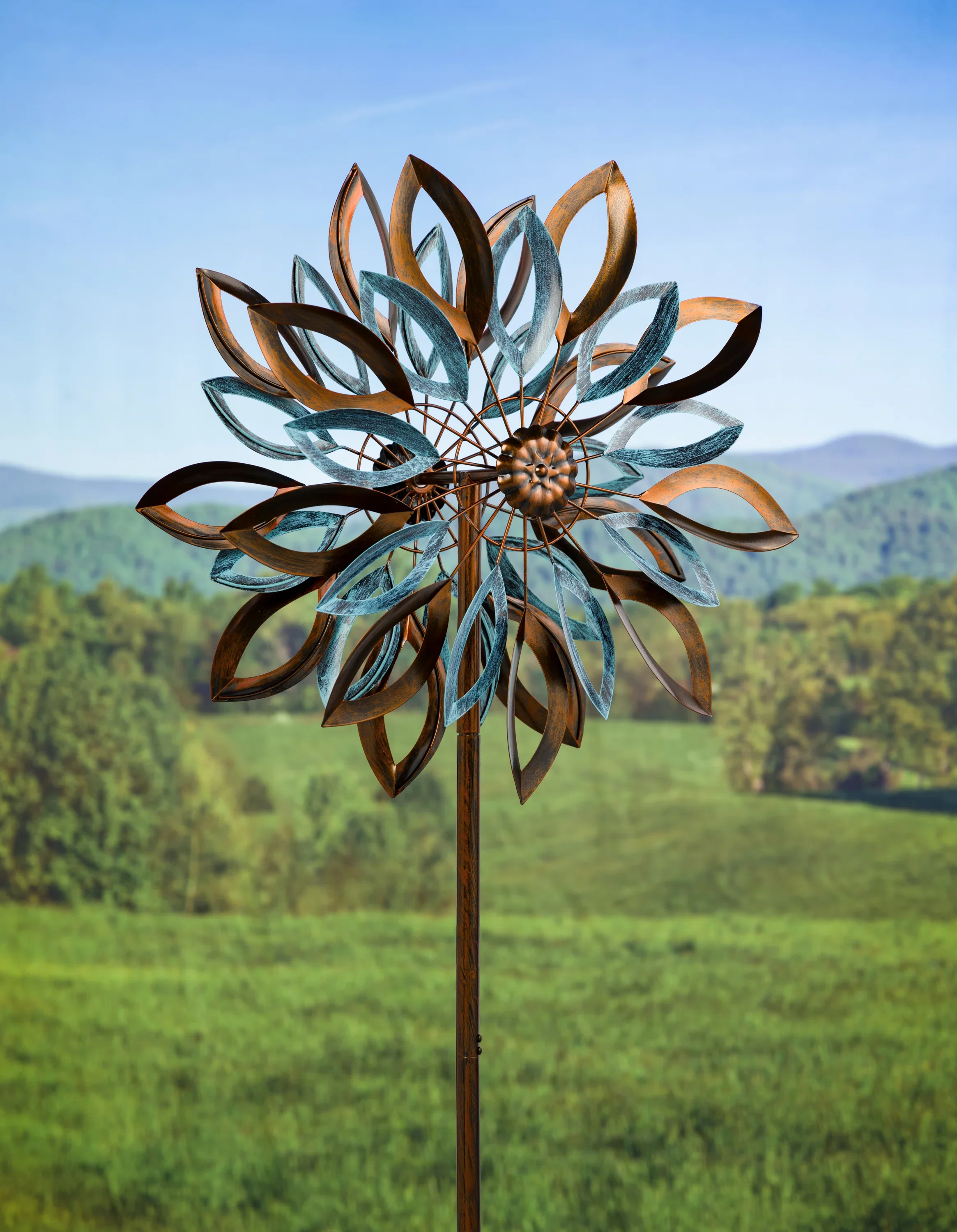 Metal garden sculpture resembling a flower with layered petals, mounted on a tall pole, against a backdrop of green hills and a clear blue sky.
