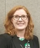 A woman with red hair, glasses, and a black blazer smiling in a professional setting.