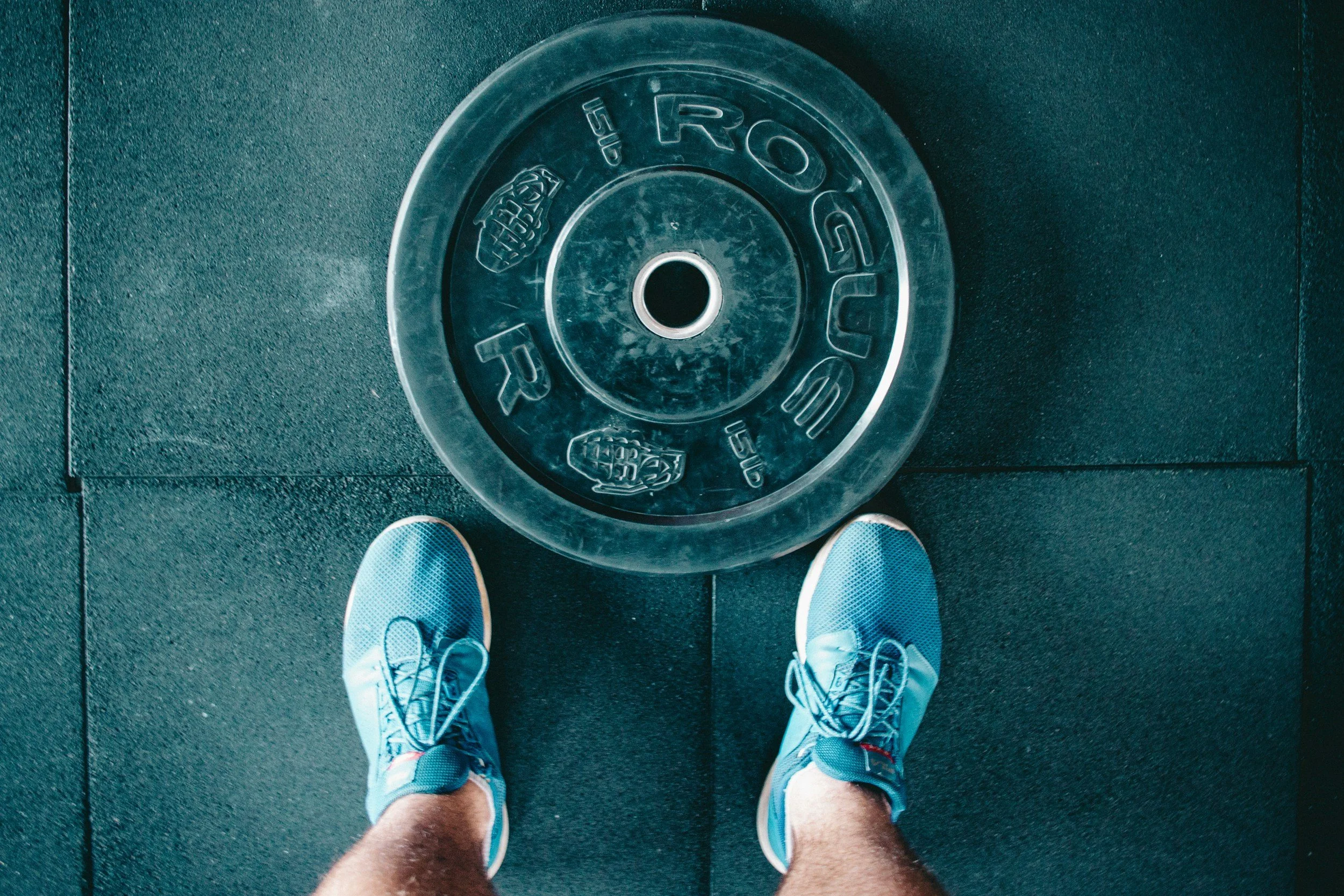 A person wearing blue sneakers standing on a gym floor, looking down at a large, round, transparent weight plate on the floor with the brand name 'Rogue' and a footprint grip design.