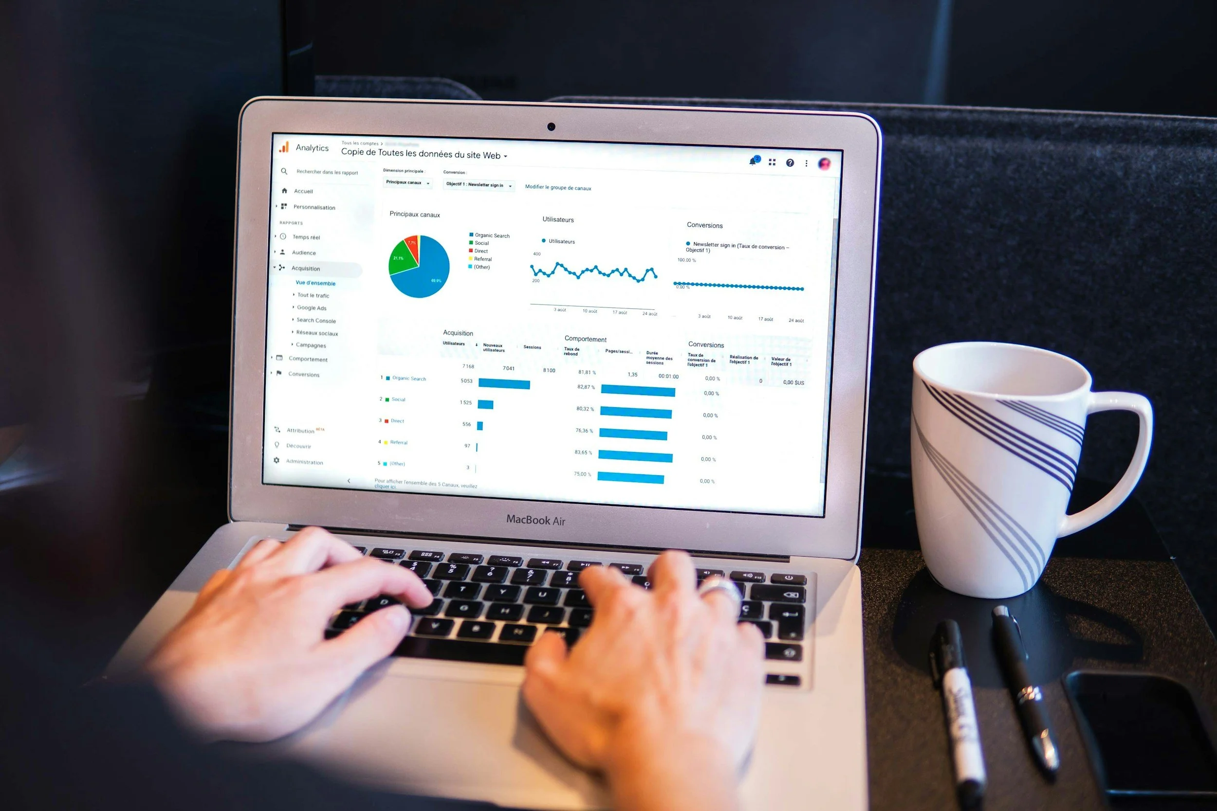 Persona usando una MacBook Air laptop con un panel de datos analíticos en la pantalla, mostrando gráficos, enseguida de una taza de cafe rayada sobre una superficie negra.
