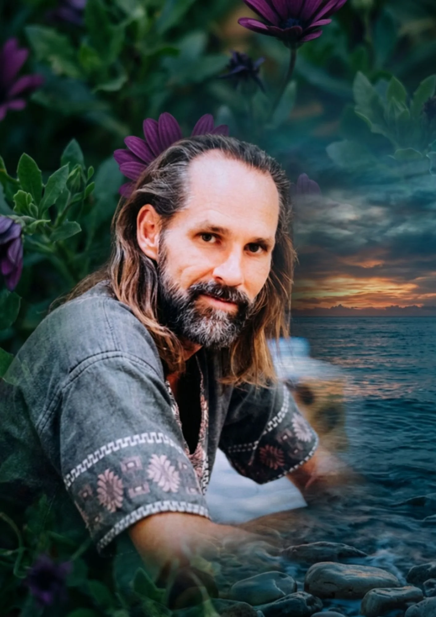 A man with long hair and a beard sitting by a body of water at sunset, surrounded by lush green plants and purple flowers.
