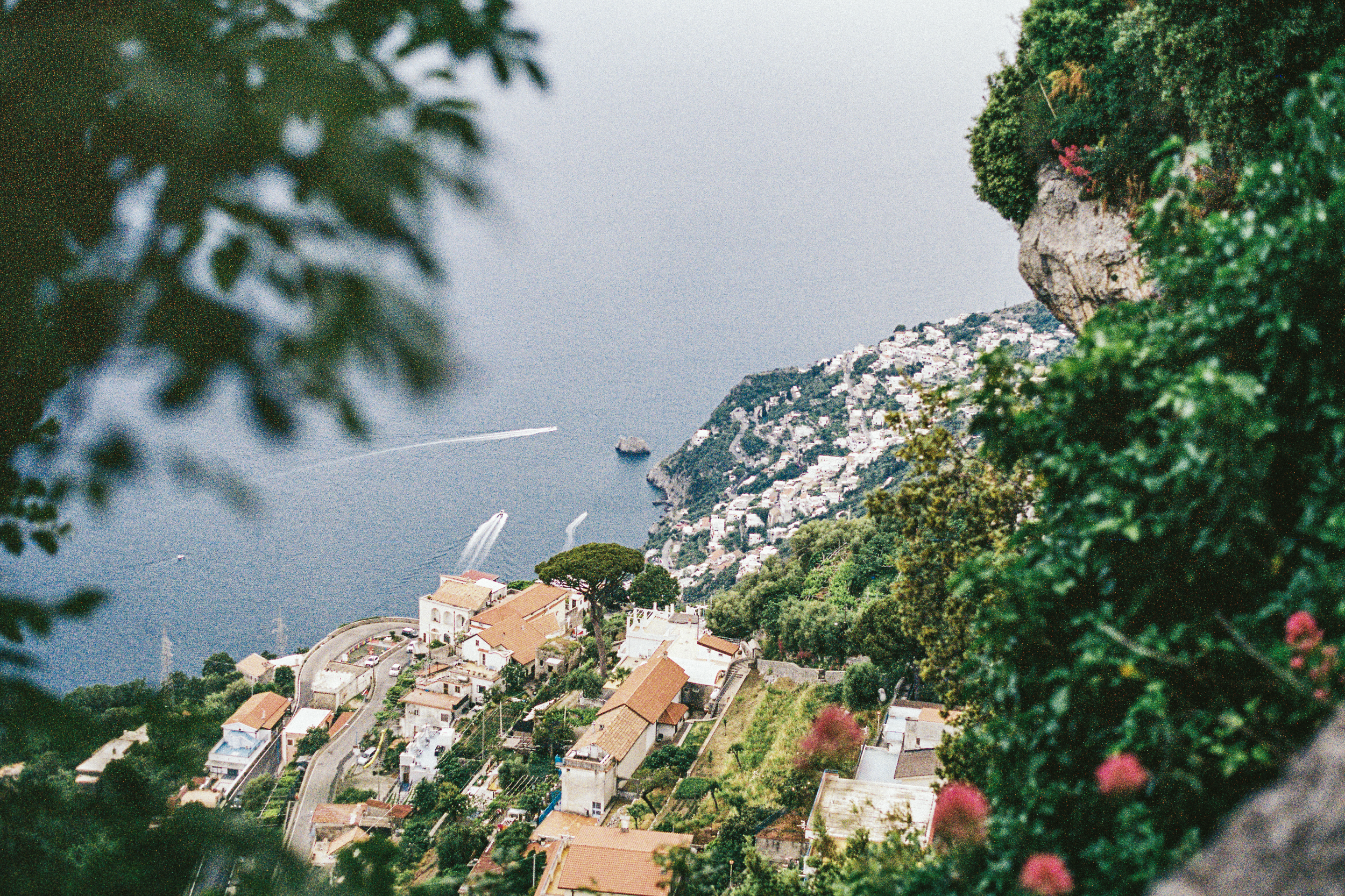 A coastal hillside town with numerous houses, a large rock formation, and a body of water with boats seen from a high vantage point surrounded by greenery.