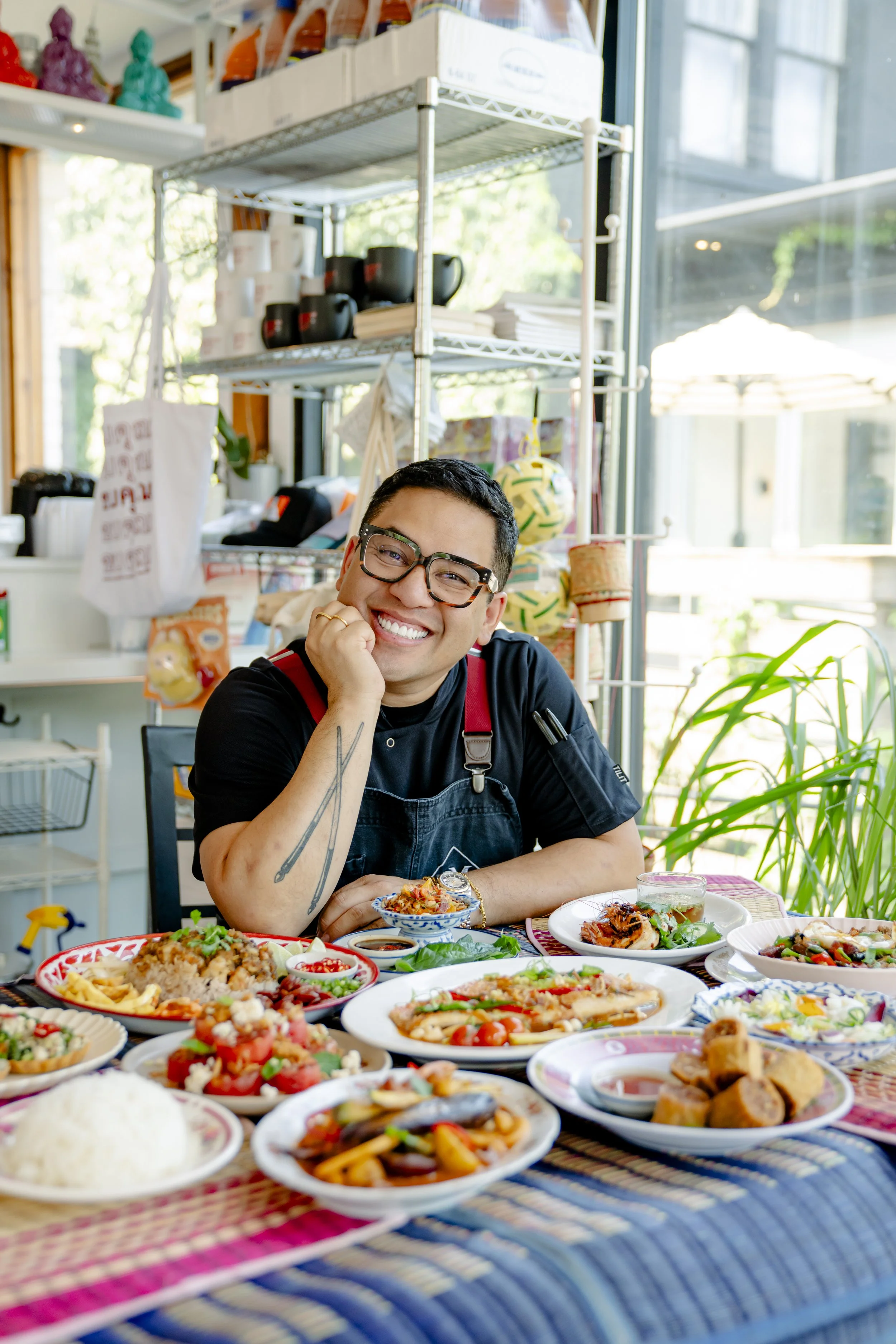A man wearing glasses and a black T-shirt sits at a table filled with colorful dishes of food, smiling. The background shows a kitchen or cafe setting with shelving containing cups, bowls, and kitchen supplies, and large windows letting in natural li