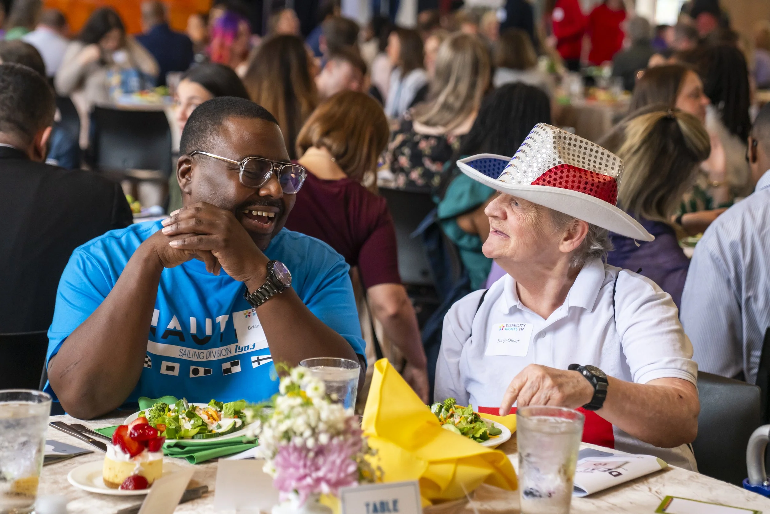 People seated at a long table enjoying a meal at a social gathering. There are visible plates of food, drinks, and festive decorations, including a woman with a white hat decorated with red, white, and silver.