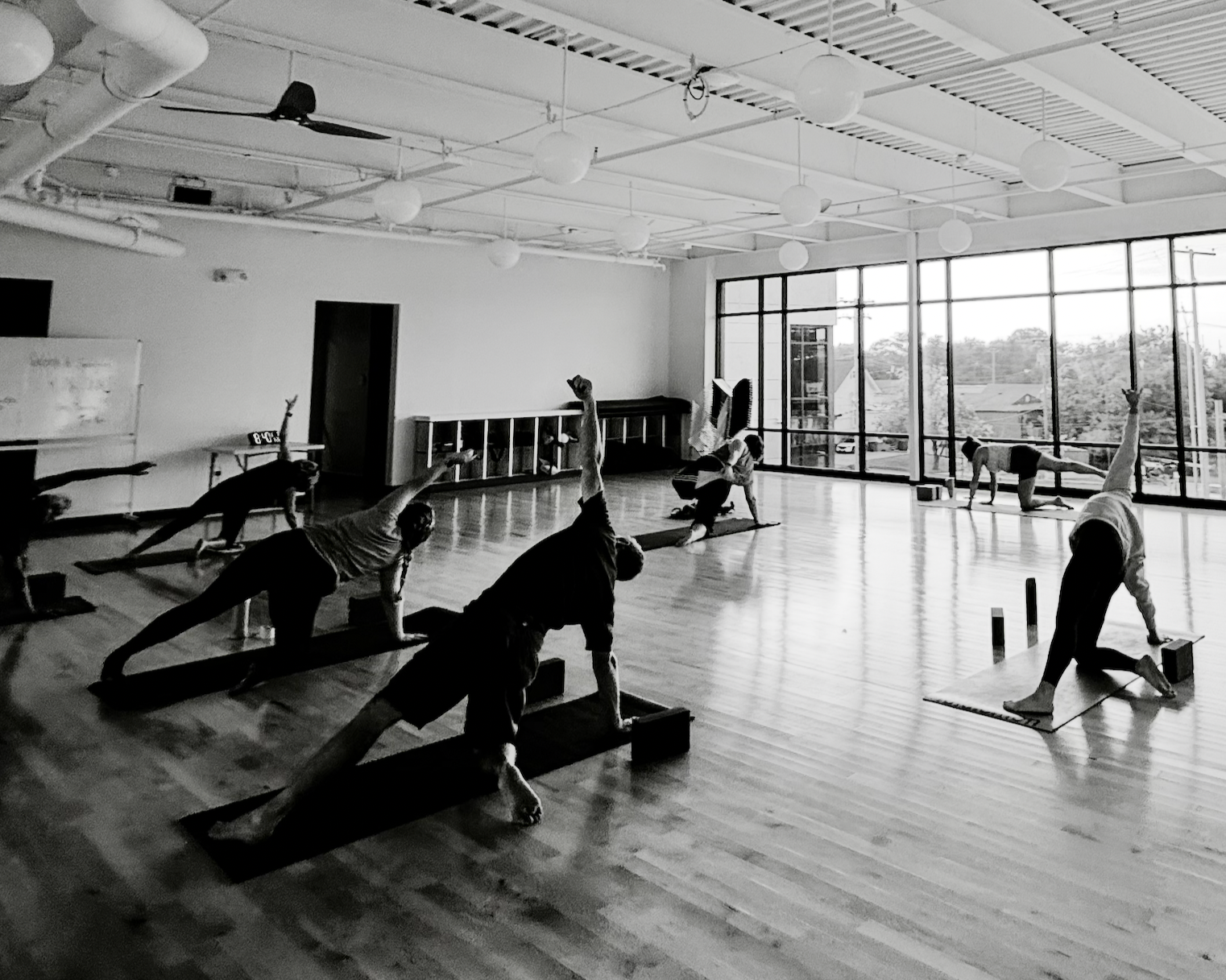 People practicing yoga in a spacious studio with large windows, wooden floors, and hanging light fixtures.