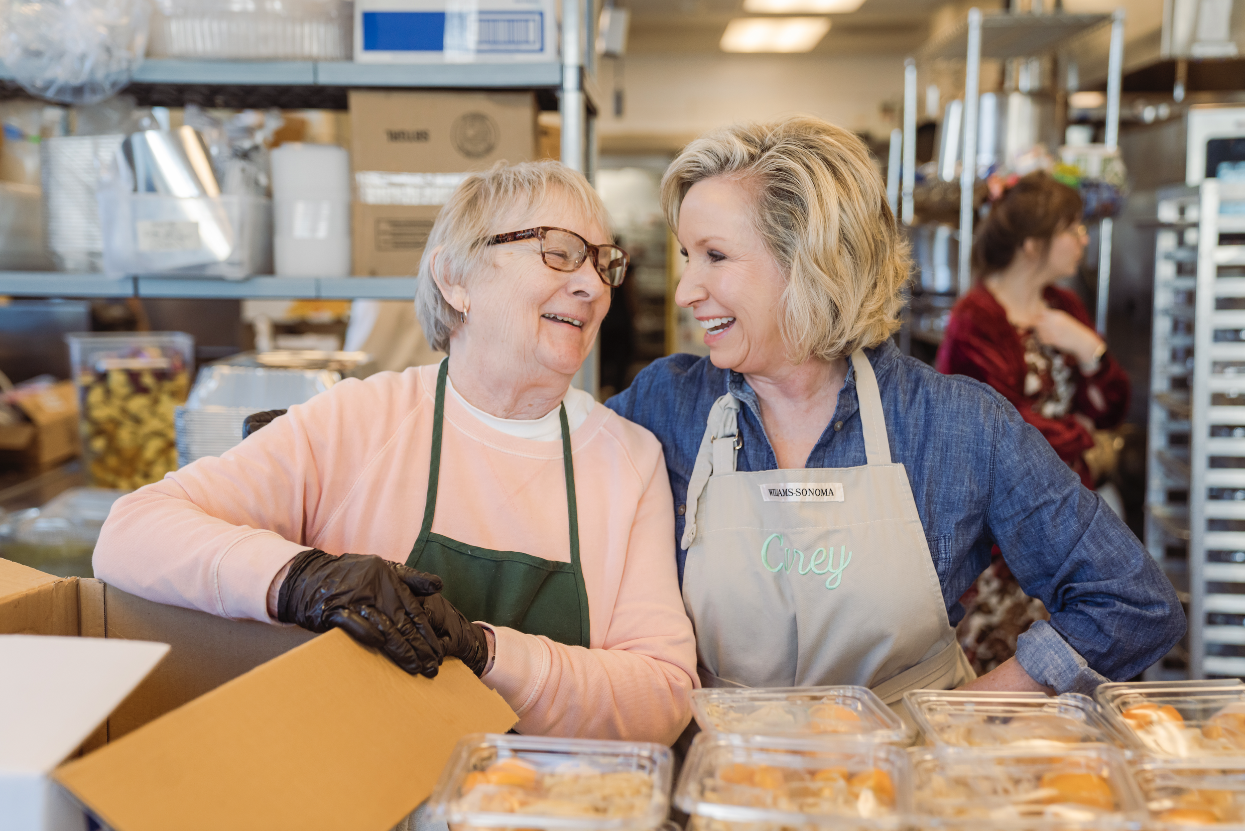 Two women smiling and working together in a food pantry or kitchen, wearing aprons. One woman has glasses and a name tag that reads 'Williams Sonoma,' while the other has a name tag that says 'Crey.' Shelves with food and supplies are in the backgrou