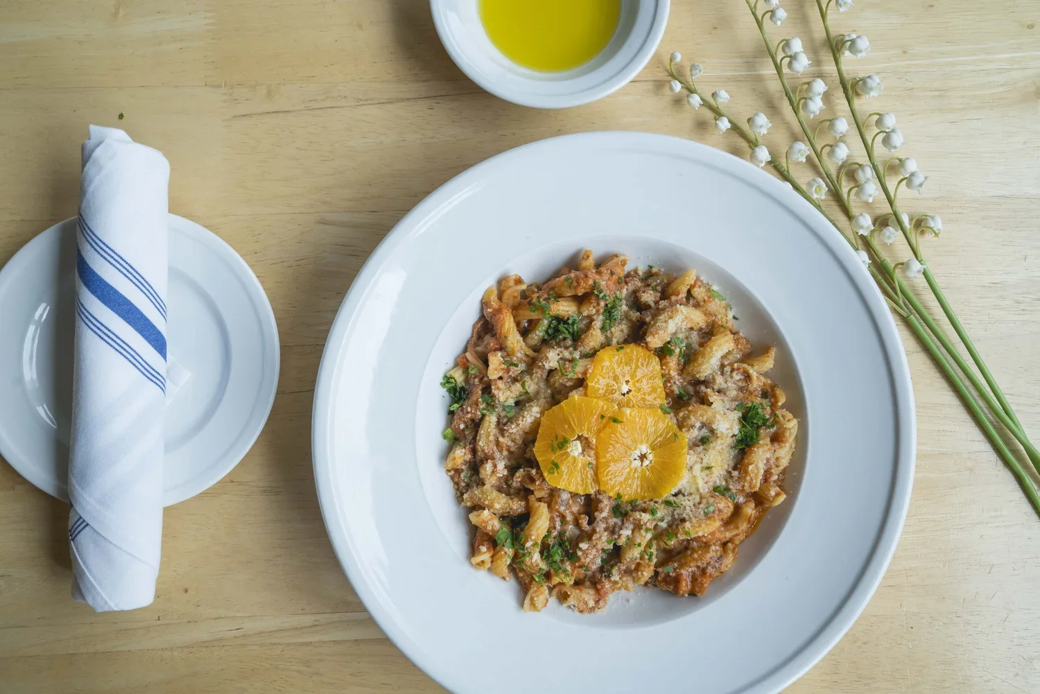 A plate of pasta with orange slices and parsley, a small dish of olive oil, a napkin with blue stripes, and a sprig of lilies of the valley on a wooden table.