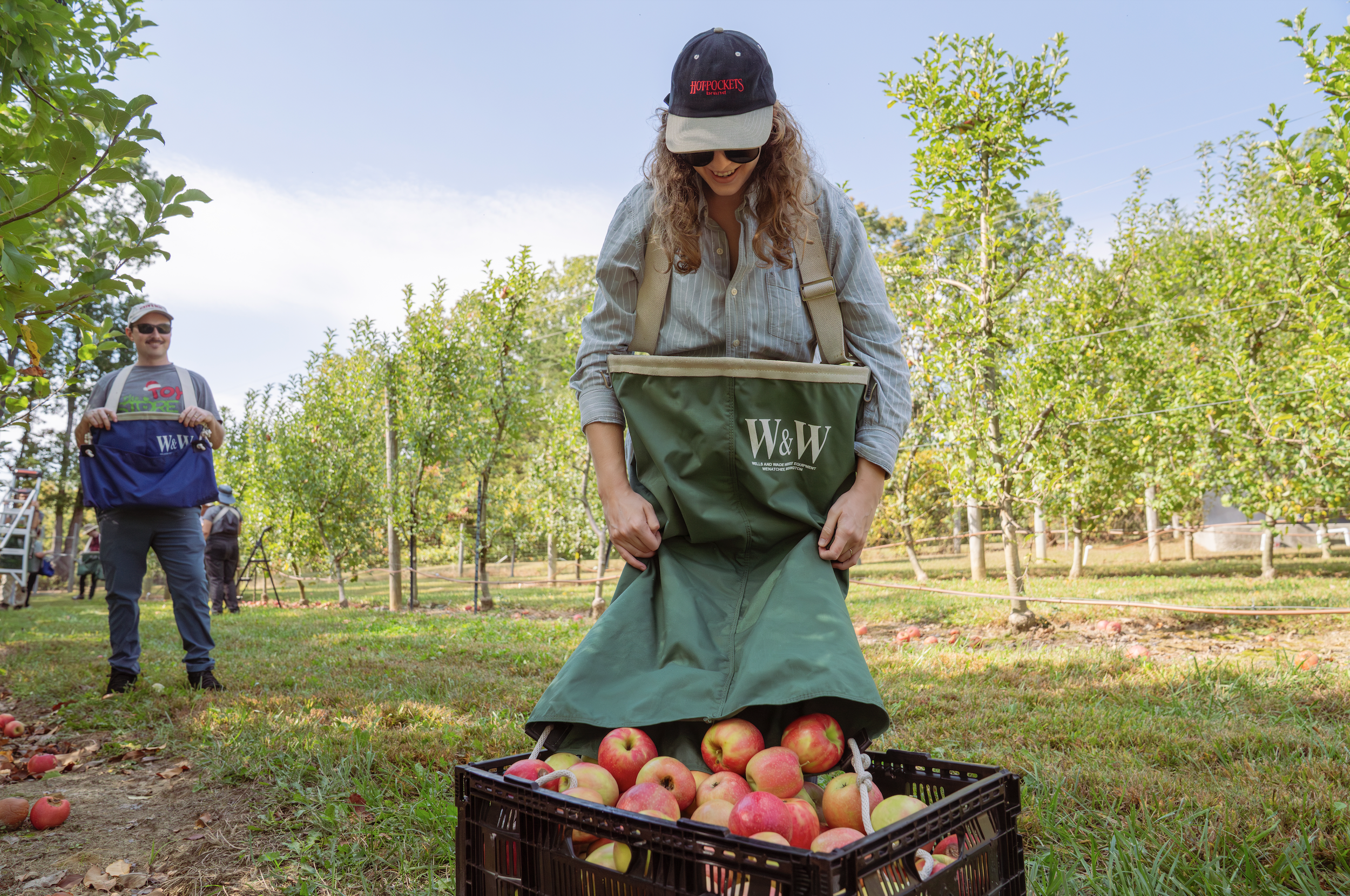 Woman in a hat and sunglasses standing in an apple orchard, collecting apples into a green bag labeled 'W&W'. A crate of apples is on the ground in front of her, and a man is visible in the background, also in the orchard.