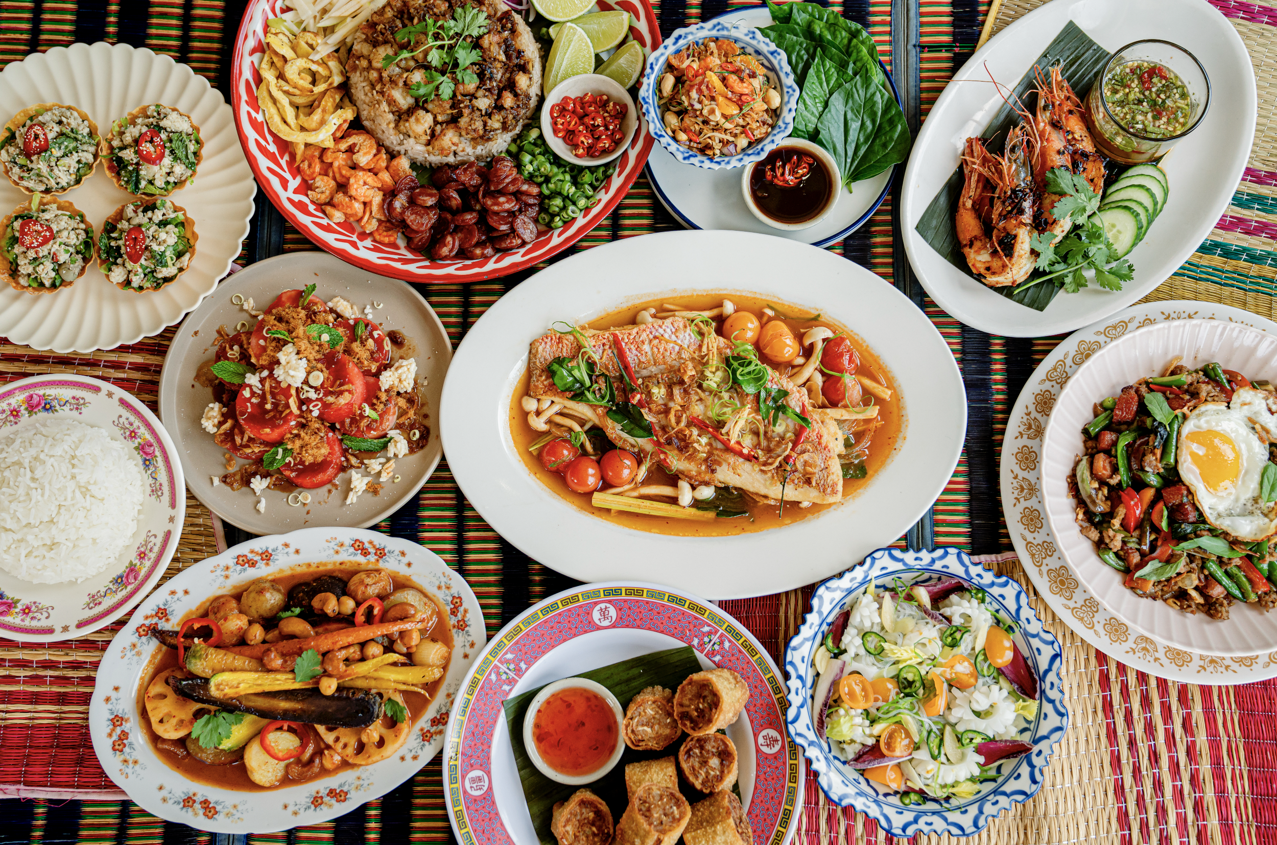 A colorful spread of various Asian dishes on woven placemats, including rice, stir-fried vegetables, noodles, grilled shrimp, mini bánh tartlets, and spring rolls.