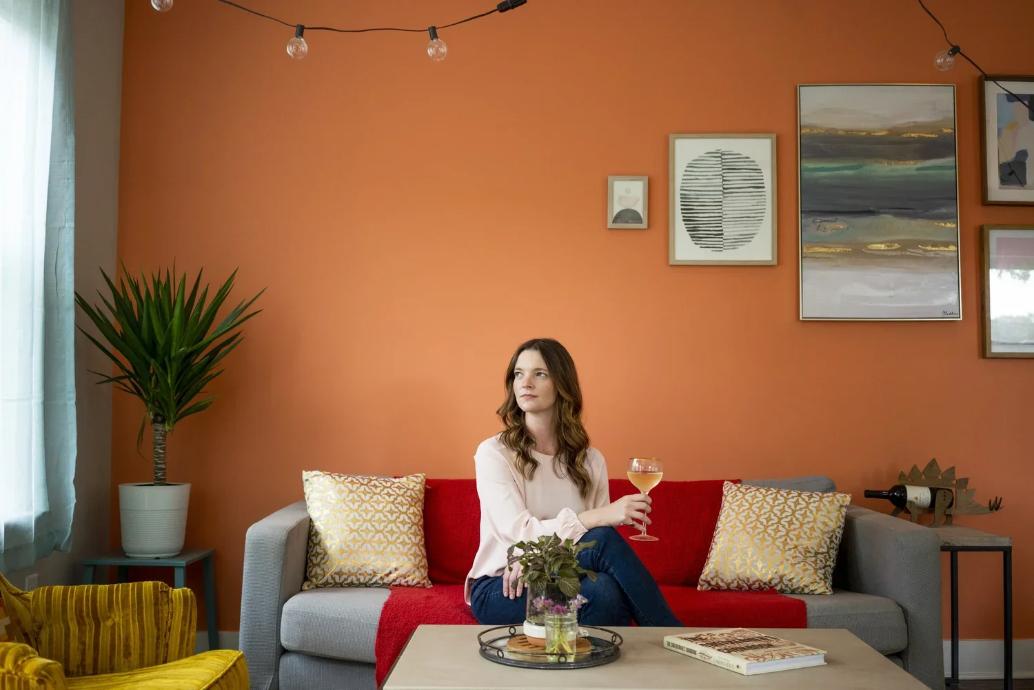 A woman sitting on a gray couch with a red blanket, holding a glass of white wine, in a living room with an orange accent wall, framed artwork, and a large potted plant, during daytime.