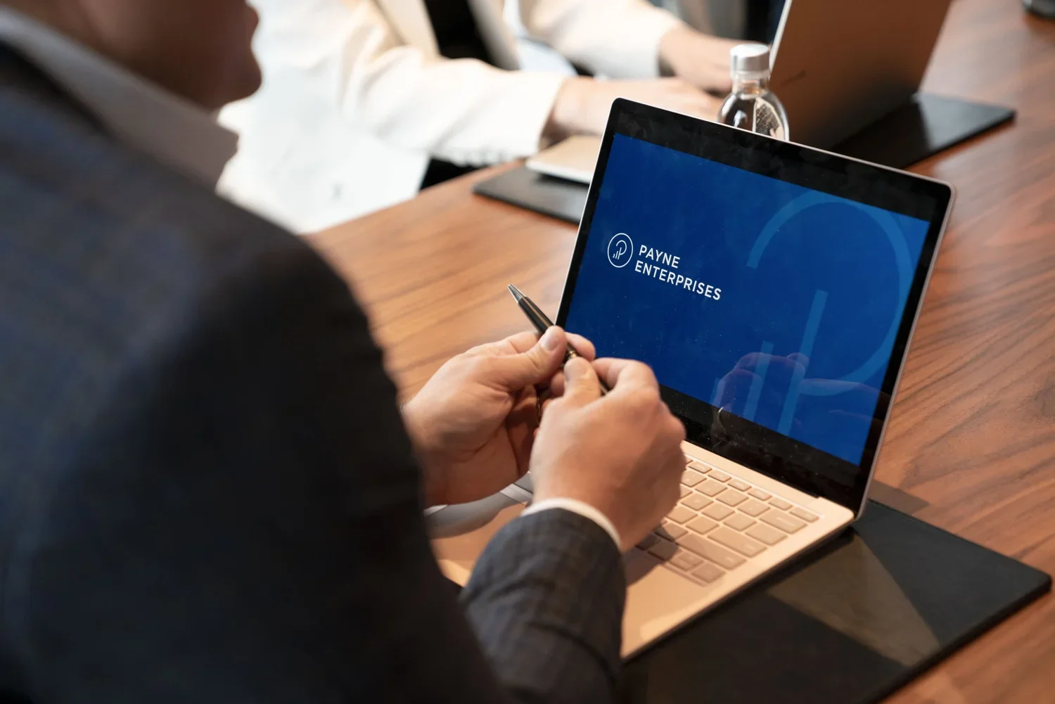 A person holding a pen, sitting at a wooden conference table with a laptop, a water bottle, and a tablet in the background. A laptop screen displays the logo and text 'PAYNE ENTERPRISES'.