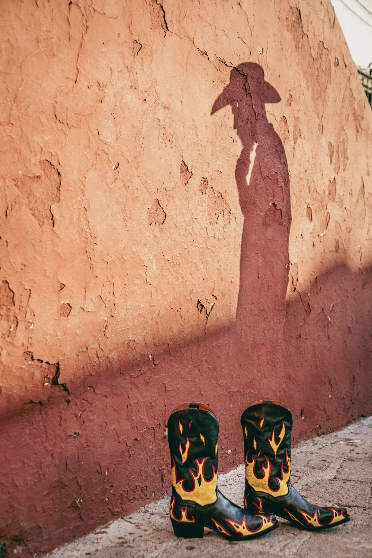 A pair of black cowboy boots with red and yellow flames design, standing on a dirt ground in front of a textured pinkish-orange wall with peeling paint.