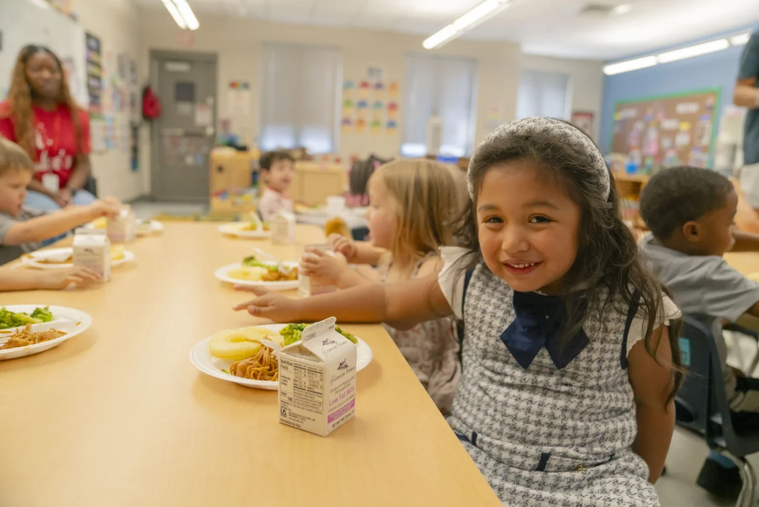 Young girl smiling and reaching for food on a cafeteria table with other children having lunch.