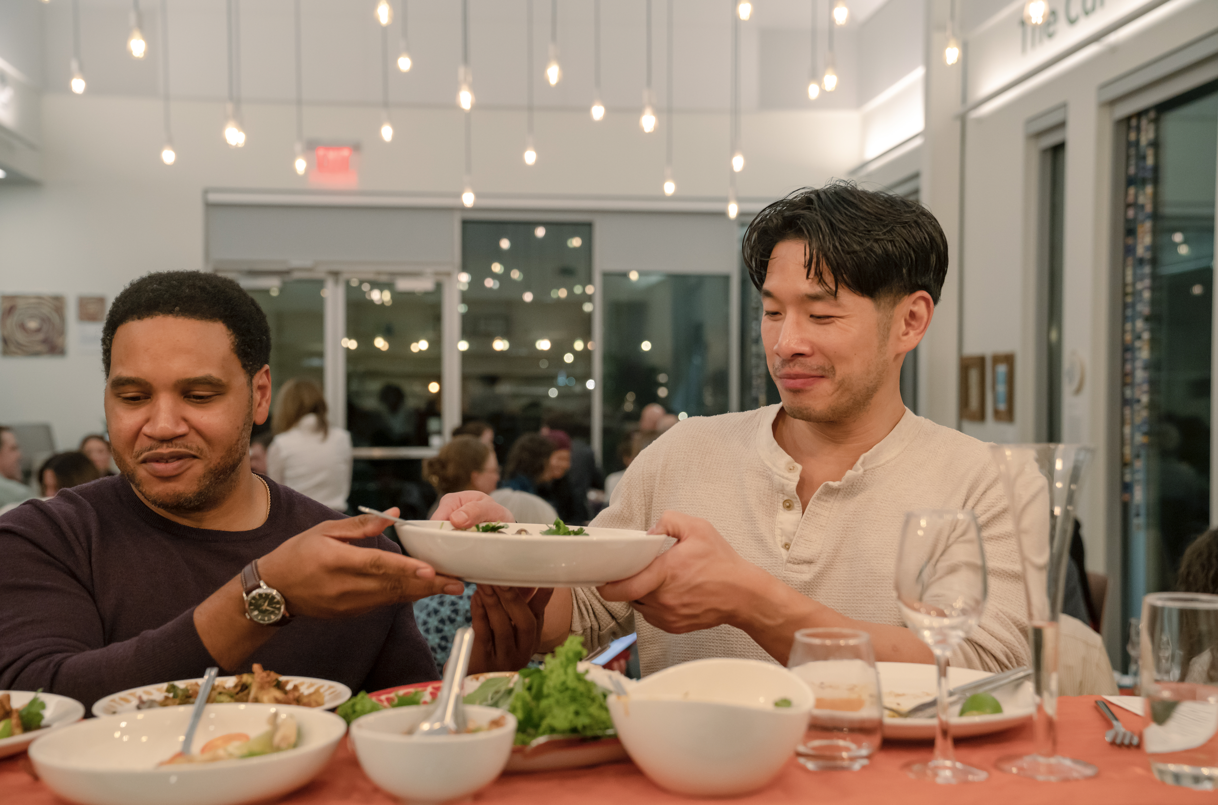 Two men sharing a bowl of food, sitting at a table with various dishes and glasses, in a well-lit restaurant with other patrons in the background.