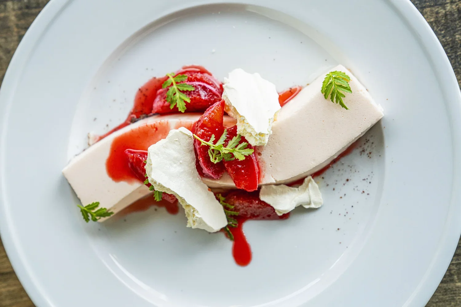 A dessert with a white mousse, topped with strawberry slices, drizzled with red berry sauce, garnished with green herbs, served on a white plate.