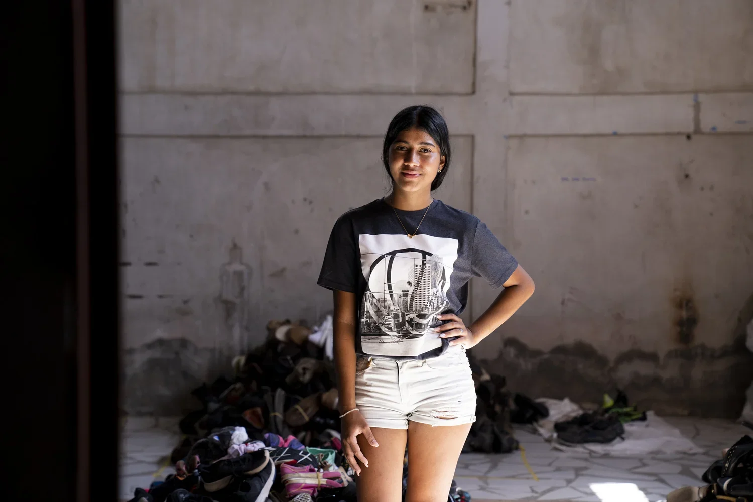 A young woman standing indoors in front of a pile of clothes and shoes, wearing a black T-shirt with a cityscape graphic and white shorts, smiling with one hand on her hip.